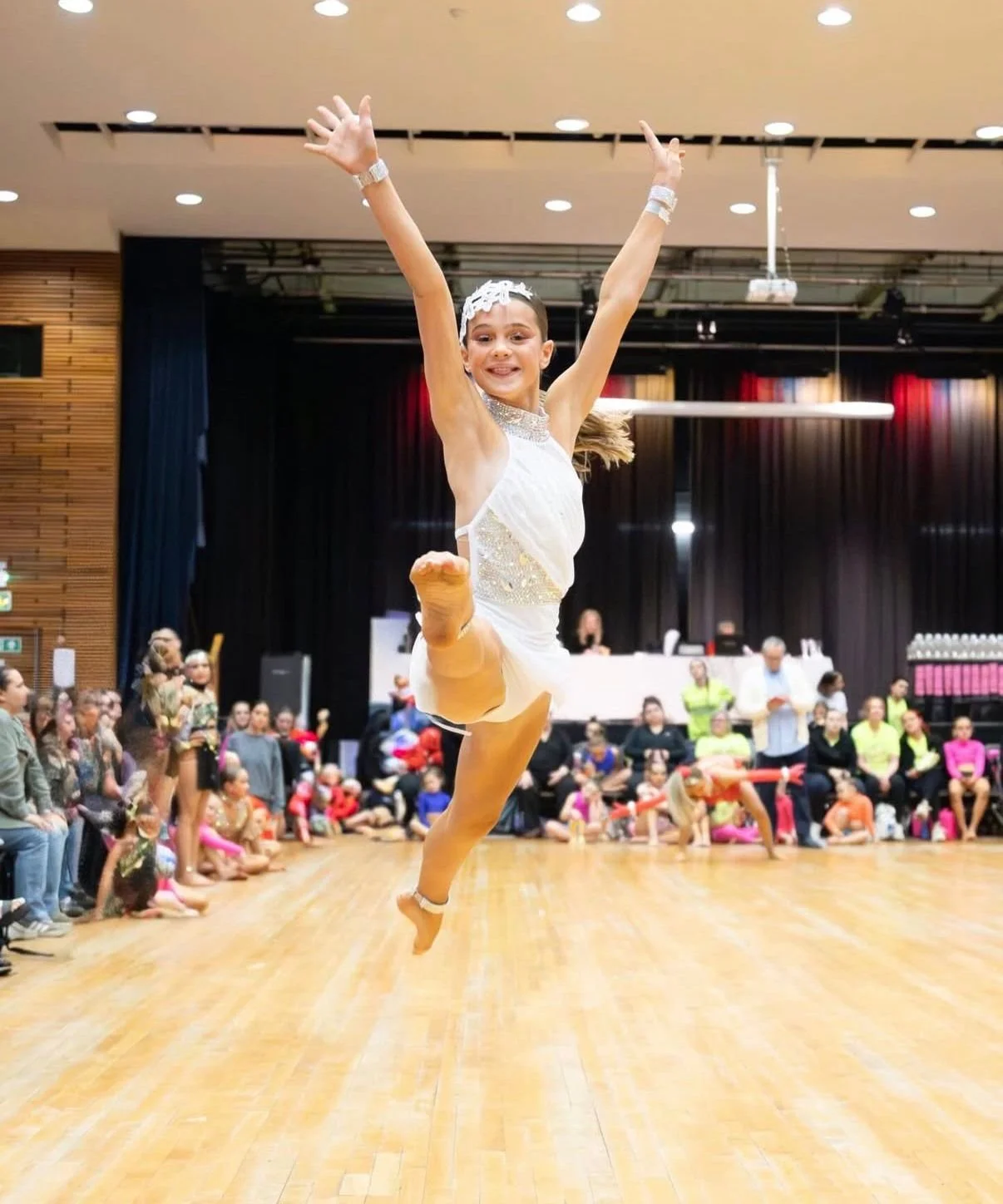 Young ballerina in a white costume performing a high kick onstage before an audience