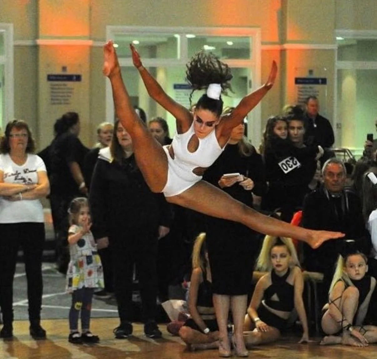 Dancer performing an aerial split in a white costume before an audience at an indoor event