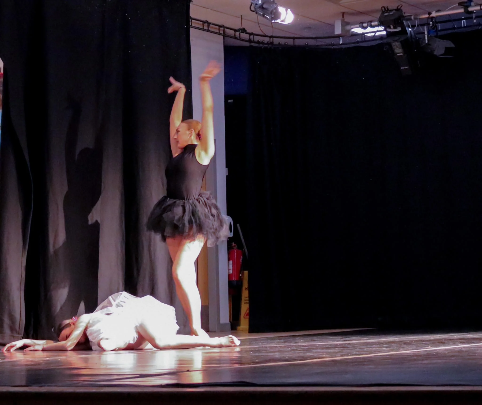 Ballet dancer performing onstage with arms raised above a dancer lying on the floor