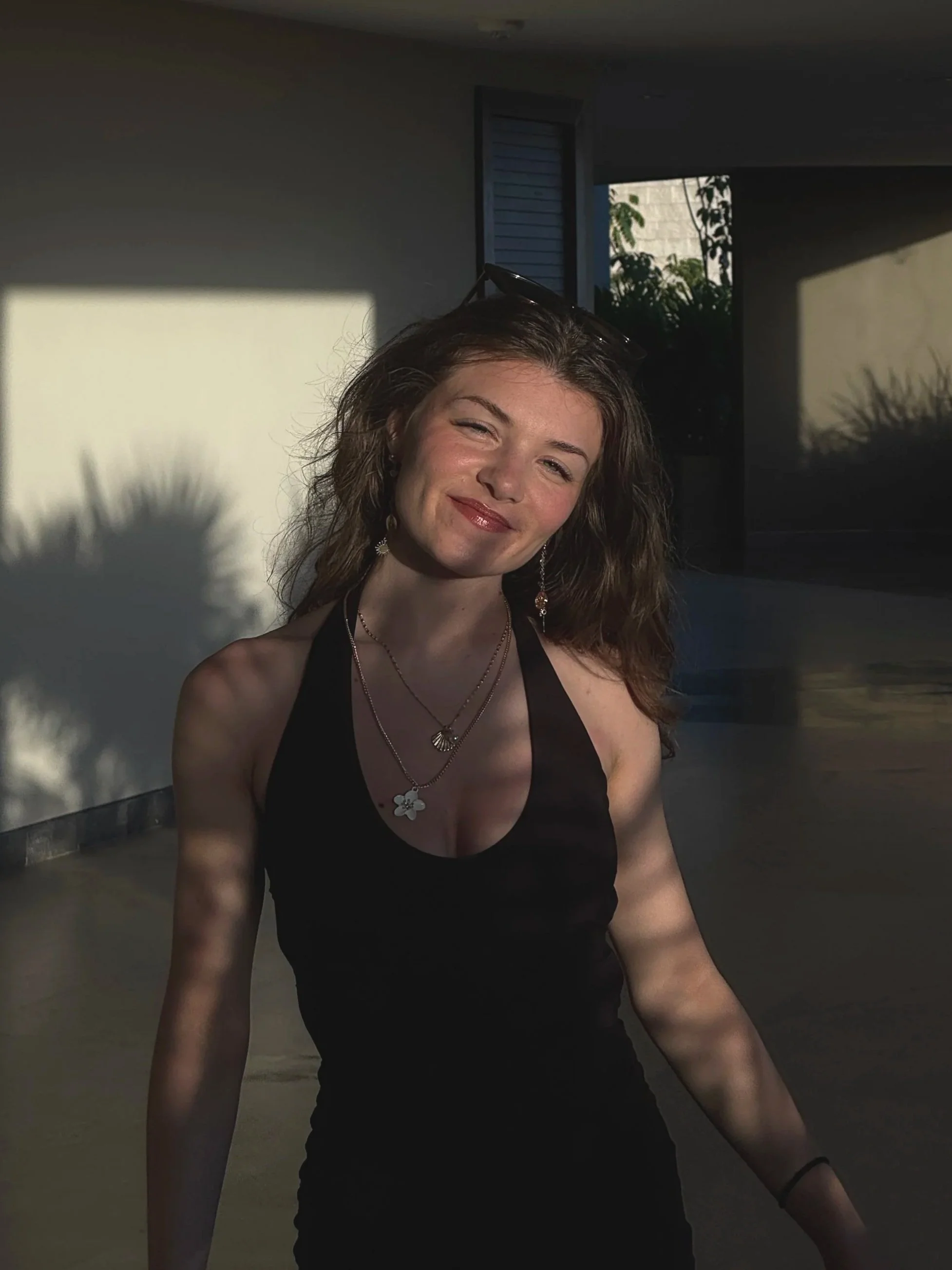 A woman with wavy brown hair, wearing a black sleeveless top and jewelry, smiling in sunlight with shadows cast on the wall behind her.