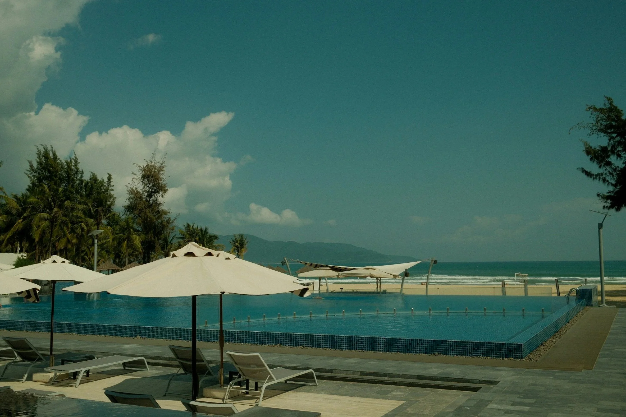 A beach scene with lounge chairs, umbrellas, a swimming pool, palm trees, and the ocean in the background under a partly cloudy sky.