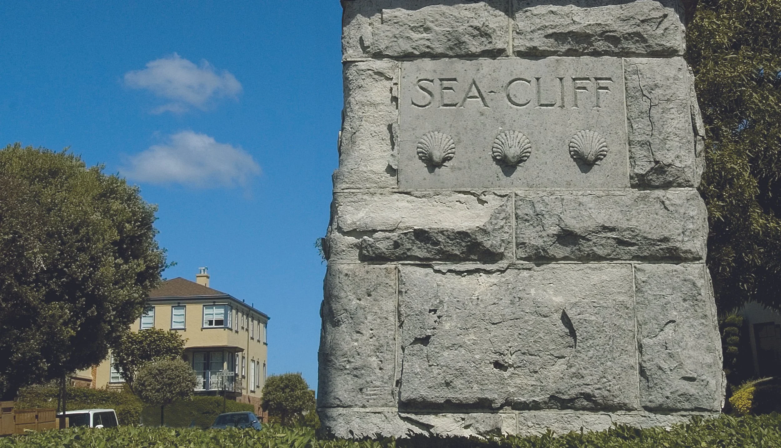 A stone sign reading 'Sea-Cliff' with three seashell carvings. In the background, there is a yellow multi-story house with white windows, trees, a parked van, and a blue sky with some clouds.