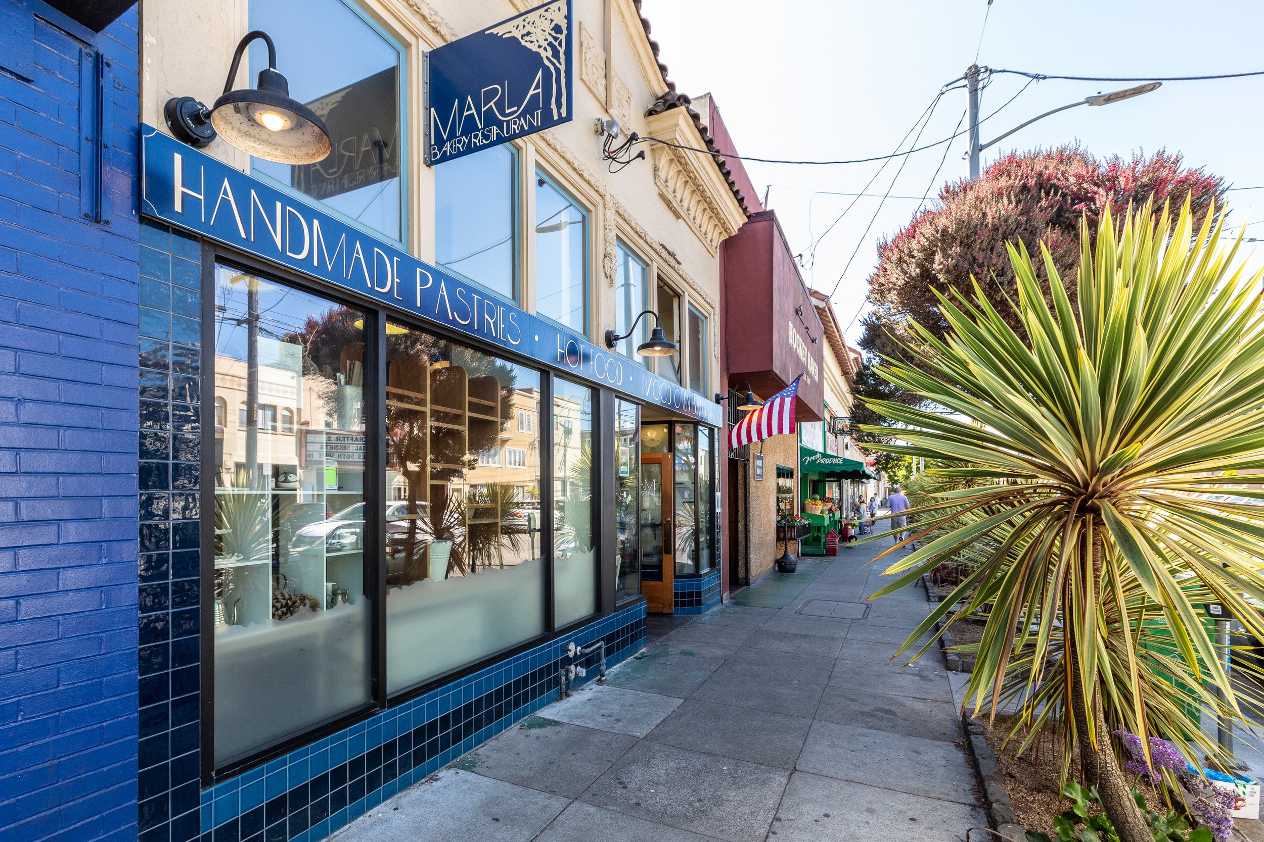 A street view with a bakery named 'Marla' on the left, featuring blue tiles and a sign advertising handmade pastries, hot food, and frozen treats. There are large windows, outdoor lighting, and a sidewalk along palm trees and colorful storefronts, wi