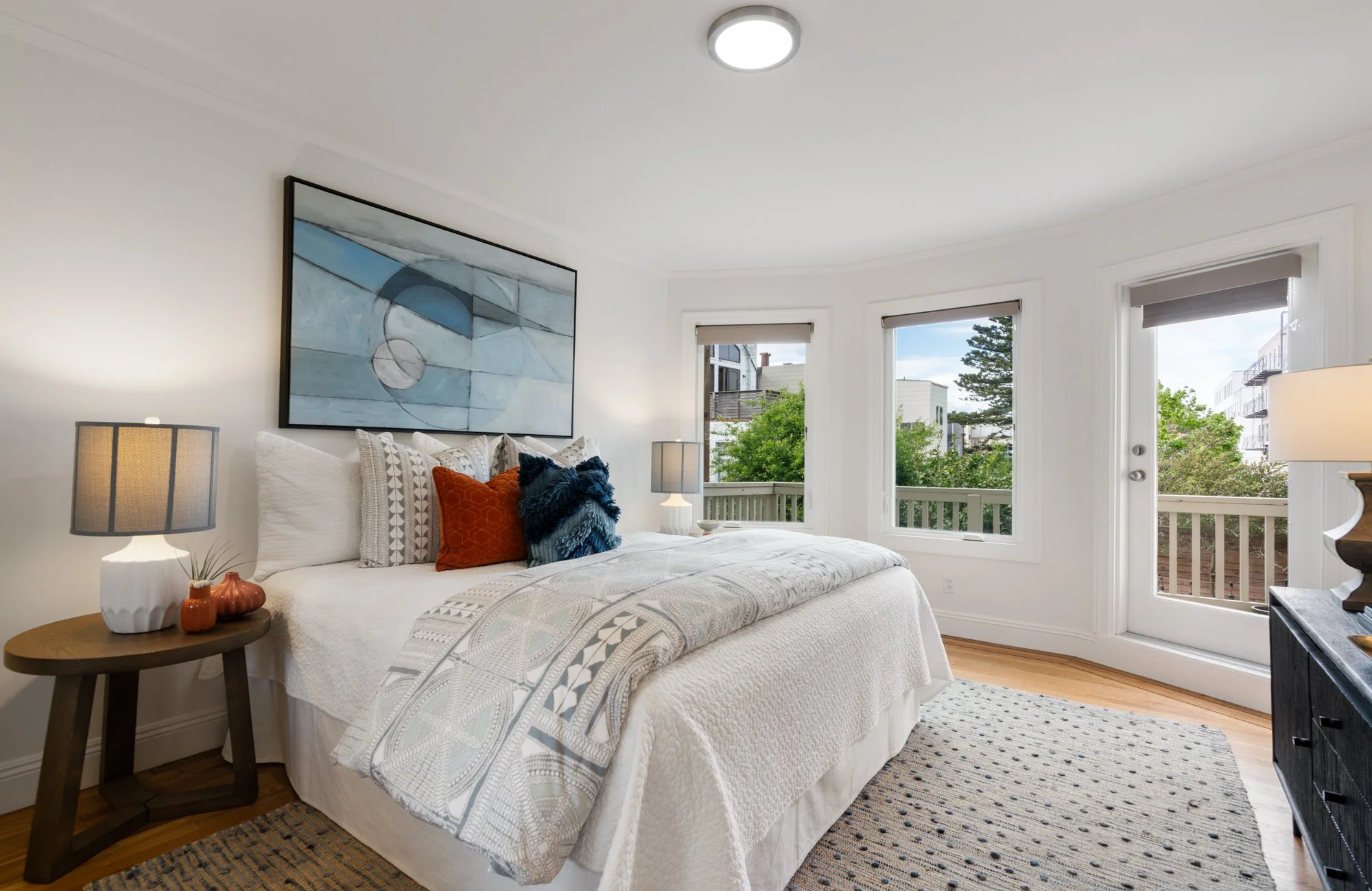 Bright bedroom with white walls and bedding, large windows showing greenery outside, modern artwork above bed, side tables with lamps, decorative pillows, and hardwood floor.