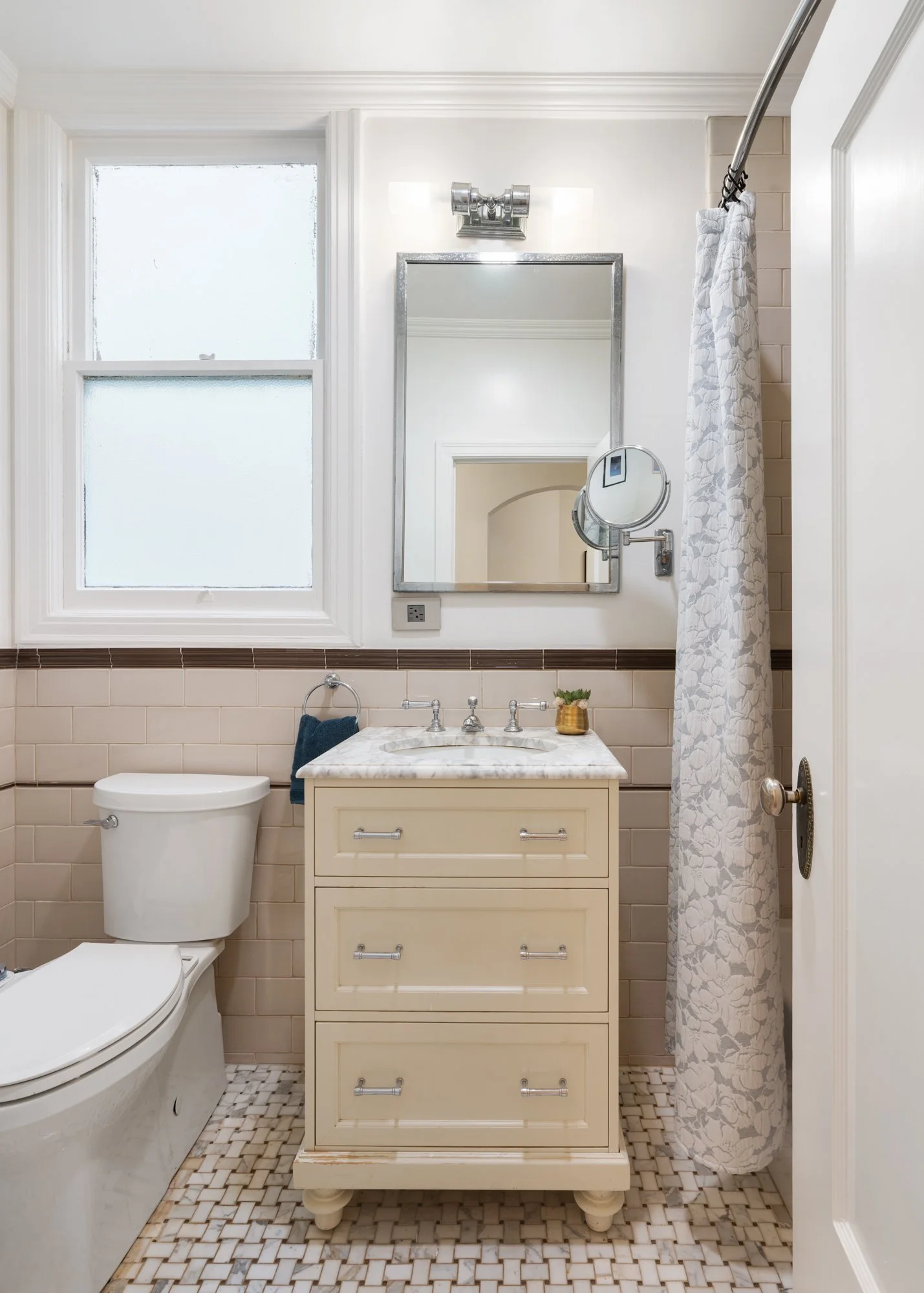 Small bathroom with beige tiled walls, white toilet, cream-colored vanity with marble top, oval mirror, window, and shower with floral-patterned curtain.