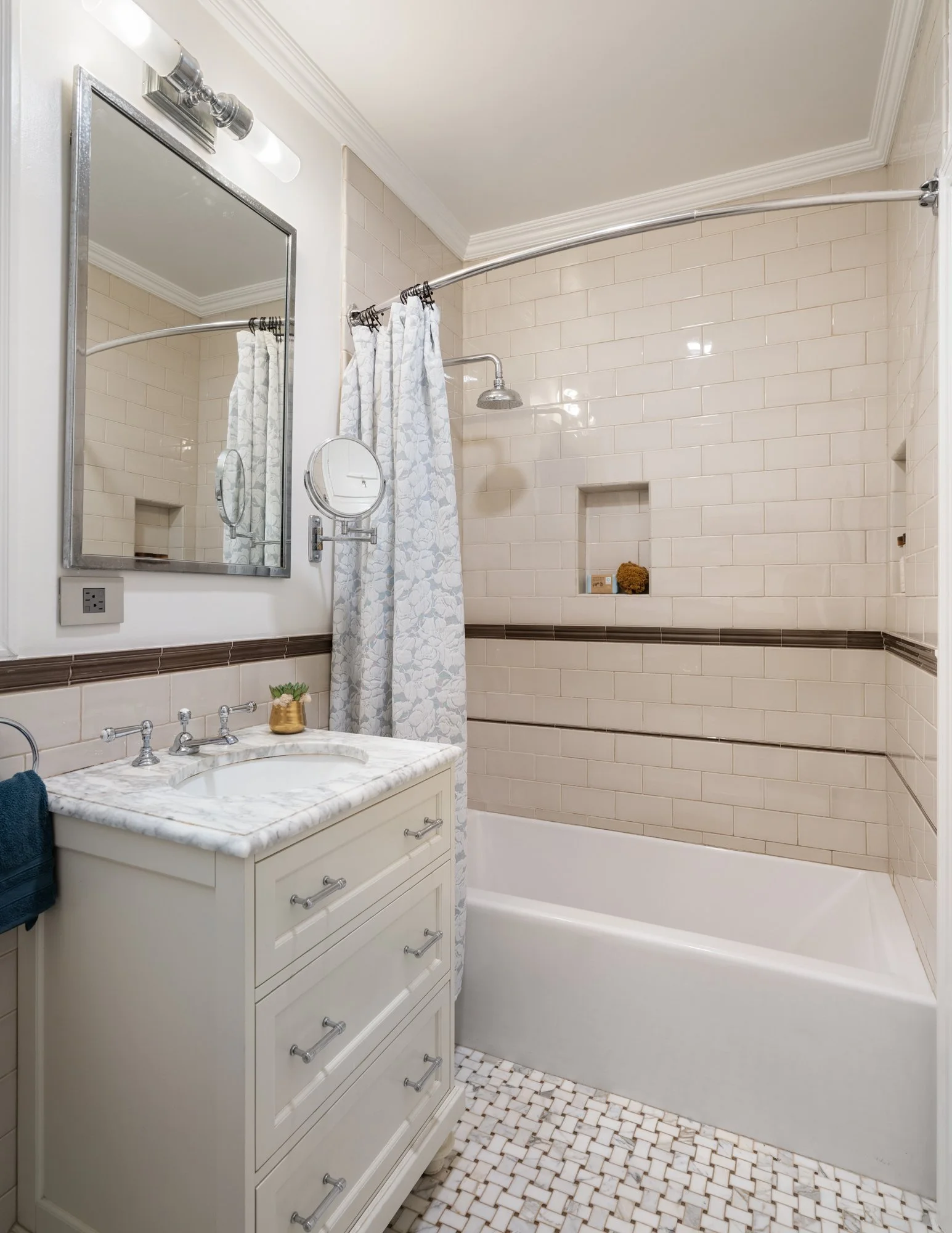 A clean and stylish bathroom with a white vanity, marble countertop, and a mirror. A shower with a curved shower rod, white tile walls, and built-in shelves. There are small decorative accents and a dark towel hanging on the side.