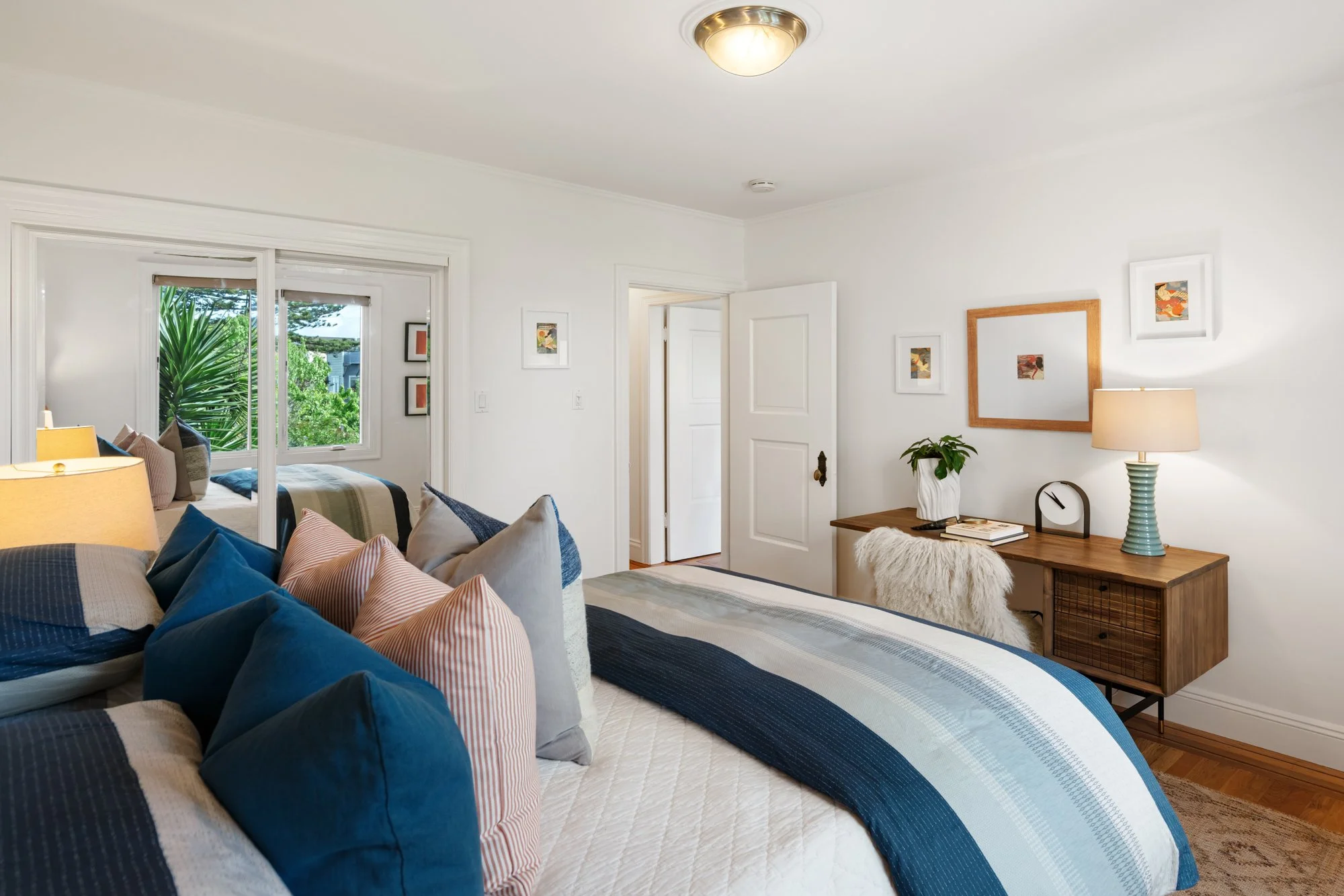 A cozy bedroom with a bed covered in blue and white striped bedding and numerous pillows. A wooden desk with a lamp, a plant, and decorative items is against the wall. The room has white walls, hardwood floors, and a window showing greenery outside.