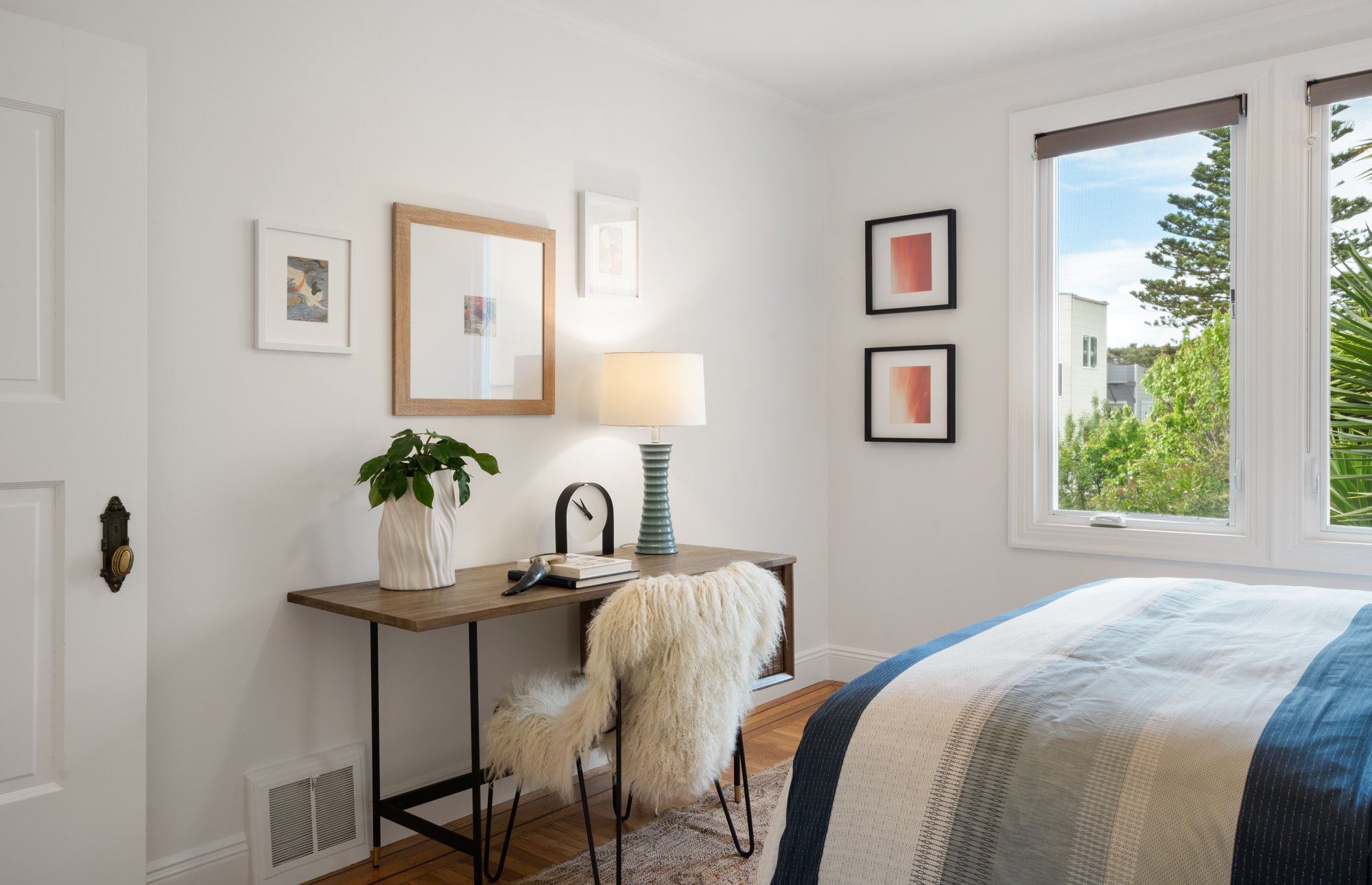 A bedroom with a wooden desk, a fluffy white chair, a table lamp, a potted plant, framed artwork on the wall, and a window showing trees outside.