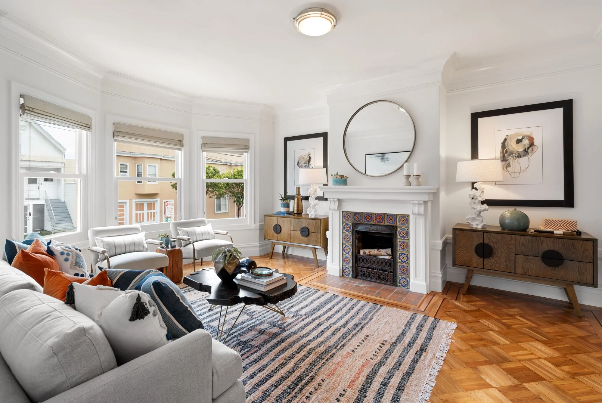 Bright living room with large windows, white sofa with colorful pillows, two white armchairs, a wooden sideboard, a decorative fireplace, a round mirror, and artwork on the walls.
