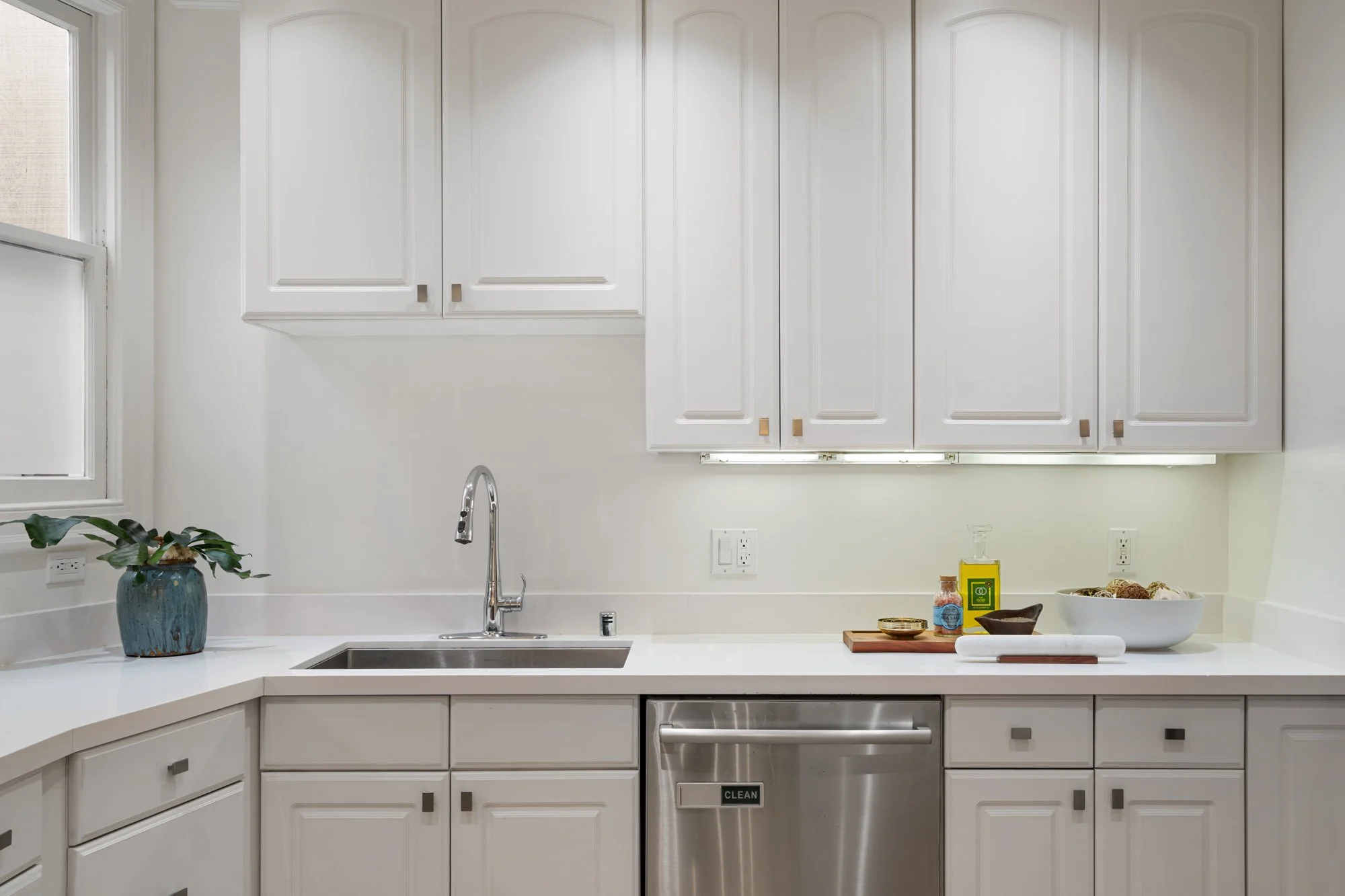 Clean, white kitchen with upper and lower cabinets, a stainless steel sink and faucet, a potted plant, and kitchen items on the counter including a bowl, a soap dispenser, and a dish towel.
