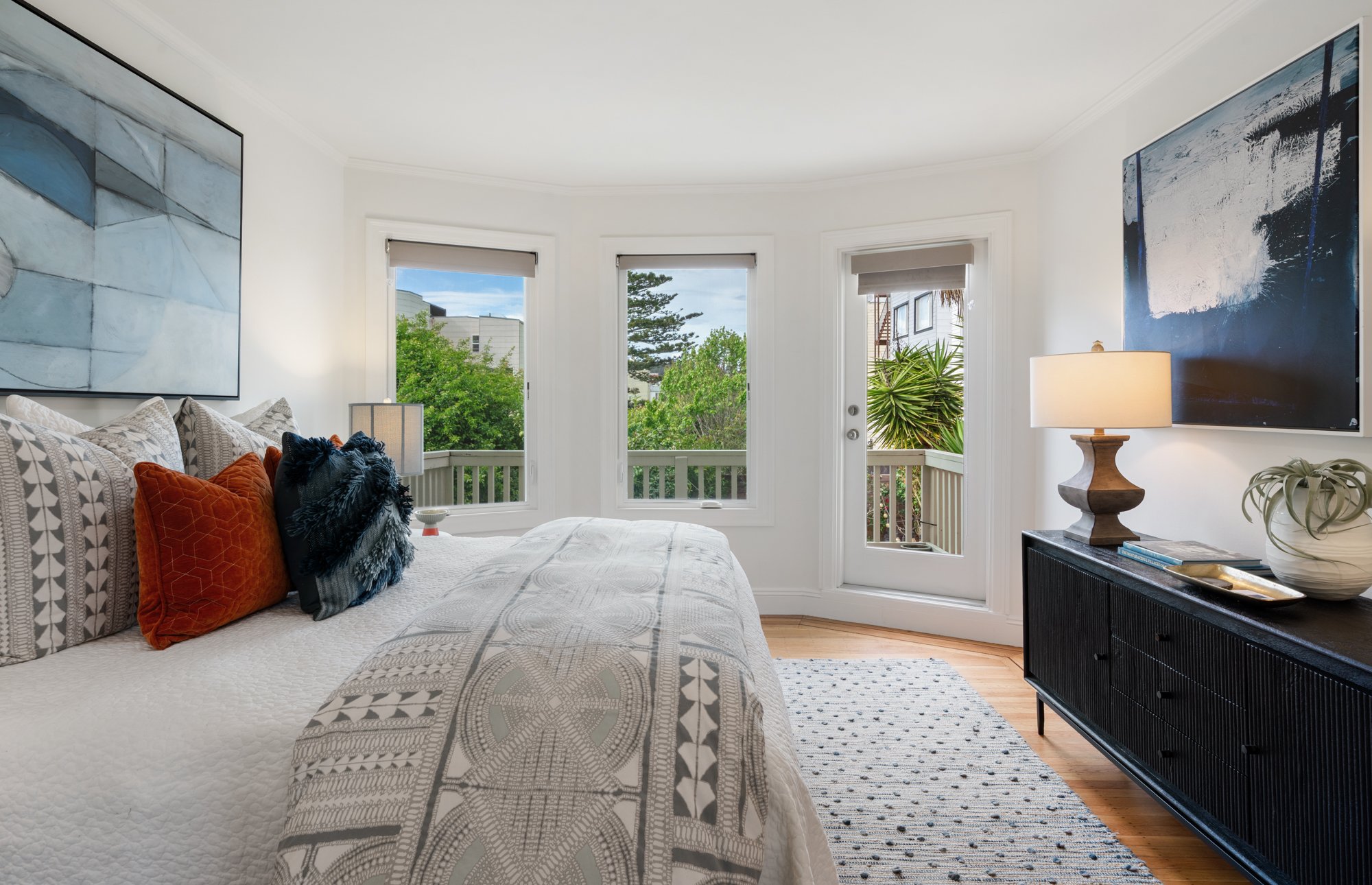 Bright bedroom with white walls, three bay windows, a door to a balcony, a large bed with decorative pillows, a black sideboard with a lamp, artwork on the walls, and a woven rug.