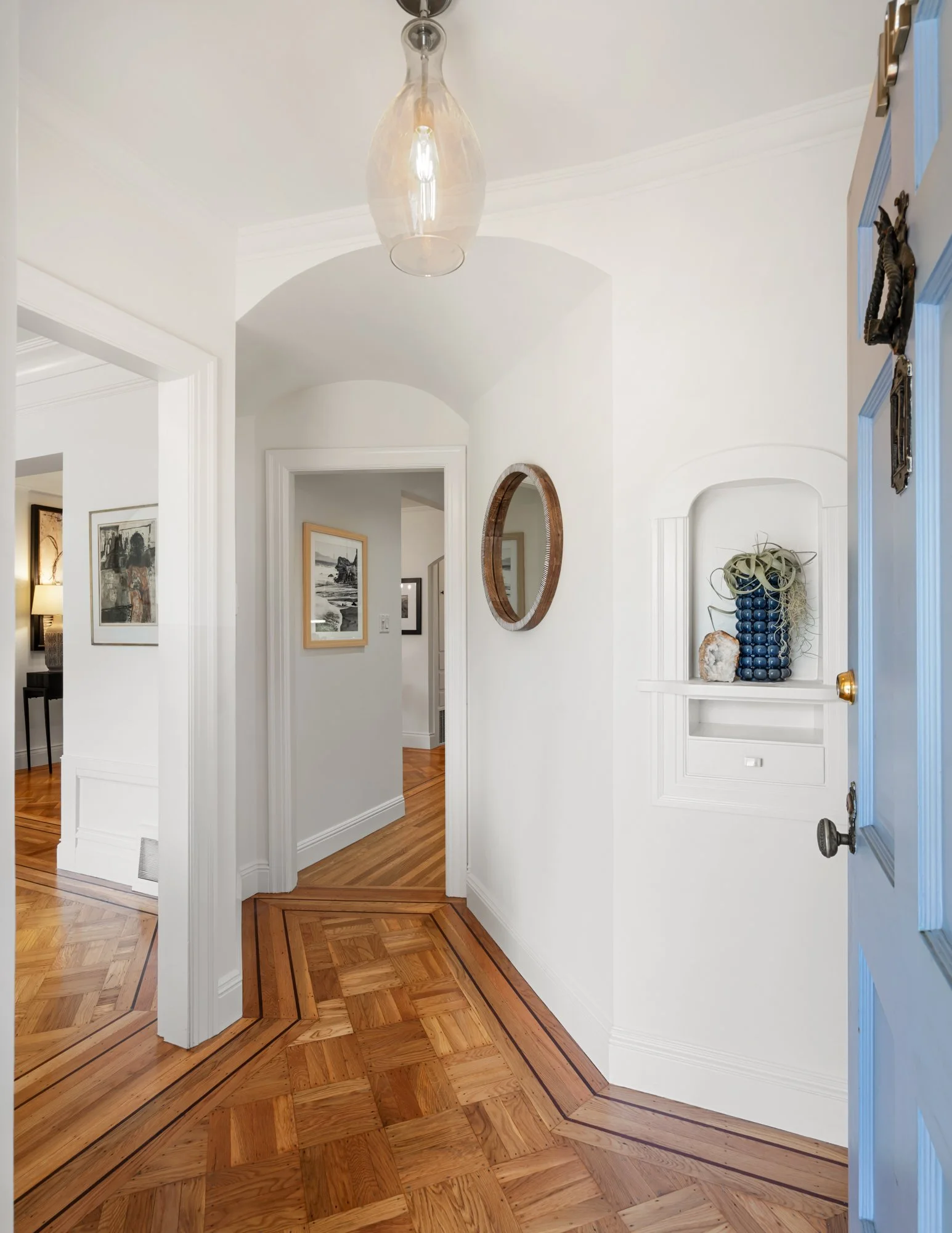 Entryway with white painted walls, wooden flooring with intricate pattern, a glass pendant light hanging from ceiling, and decorative mirrors and a small niche with potpourri and a decorative rock.