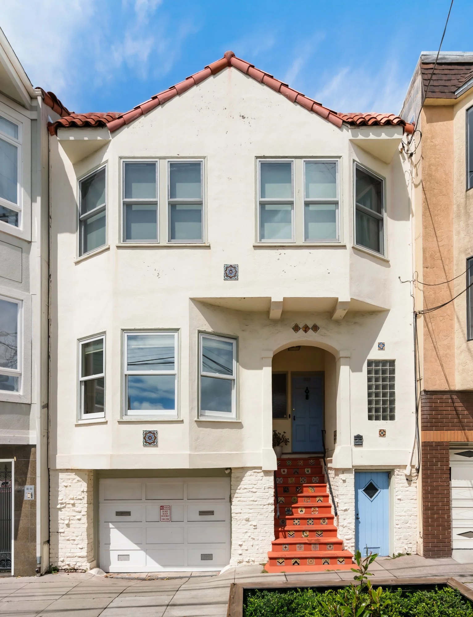 A white multi-story house with a red-tiled gable roof, featuring several windows with clear glass panes, a blue front door accessed by red decorative tiled stairs, and a small garage door on the ground level. The house has some decorative tiles and a