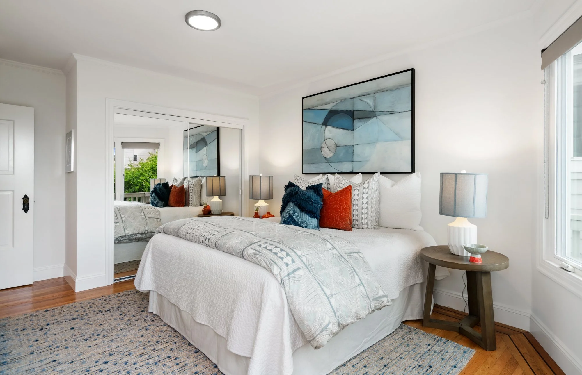 Modern bedroom with white bedding, decorative pillows in blue and orange, artwork above bed, mirrored closet doors, and a window with blinds.