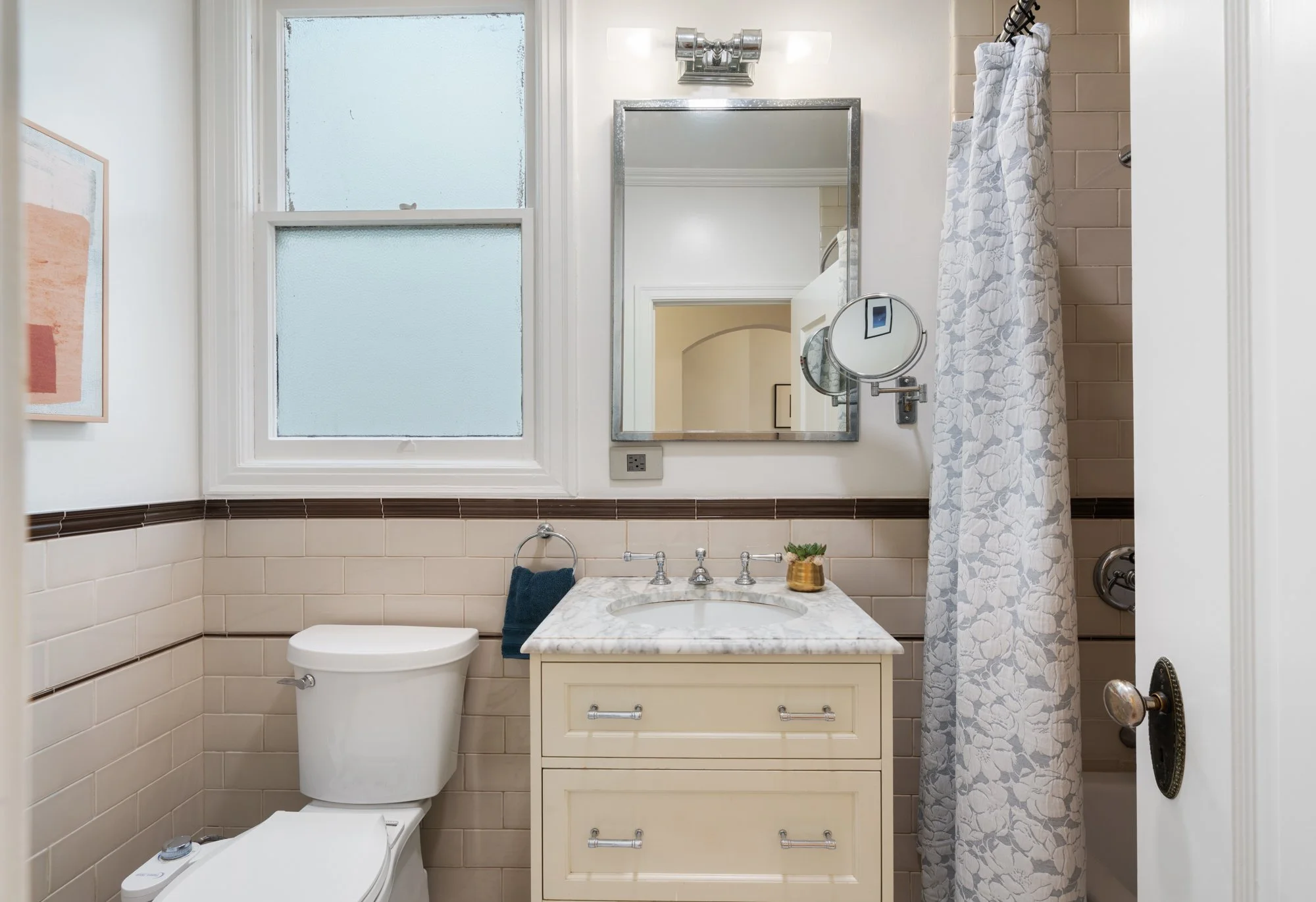Bathroom with beige tiled walls, a frosted window, a white toilet, a cream-colored vanity with a marble top, a mirror, and a shower with a floral curtain.