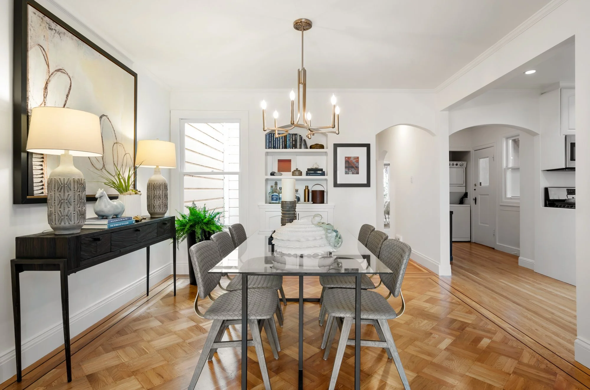 Dining room with a glass-top table, six upholstered chairs, a black sideboard with lamps and decorative items, a chandelier, and a built-in bookshelf with plants and artwork, with hardwood floors and white walls.