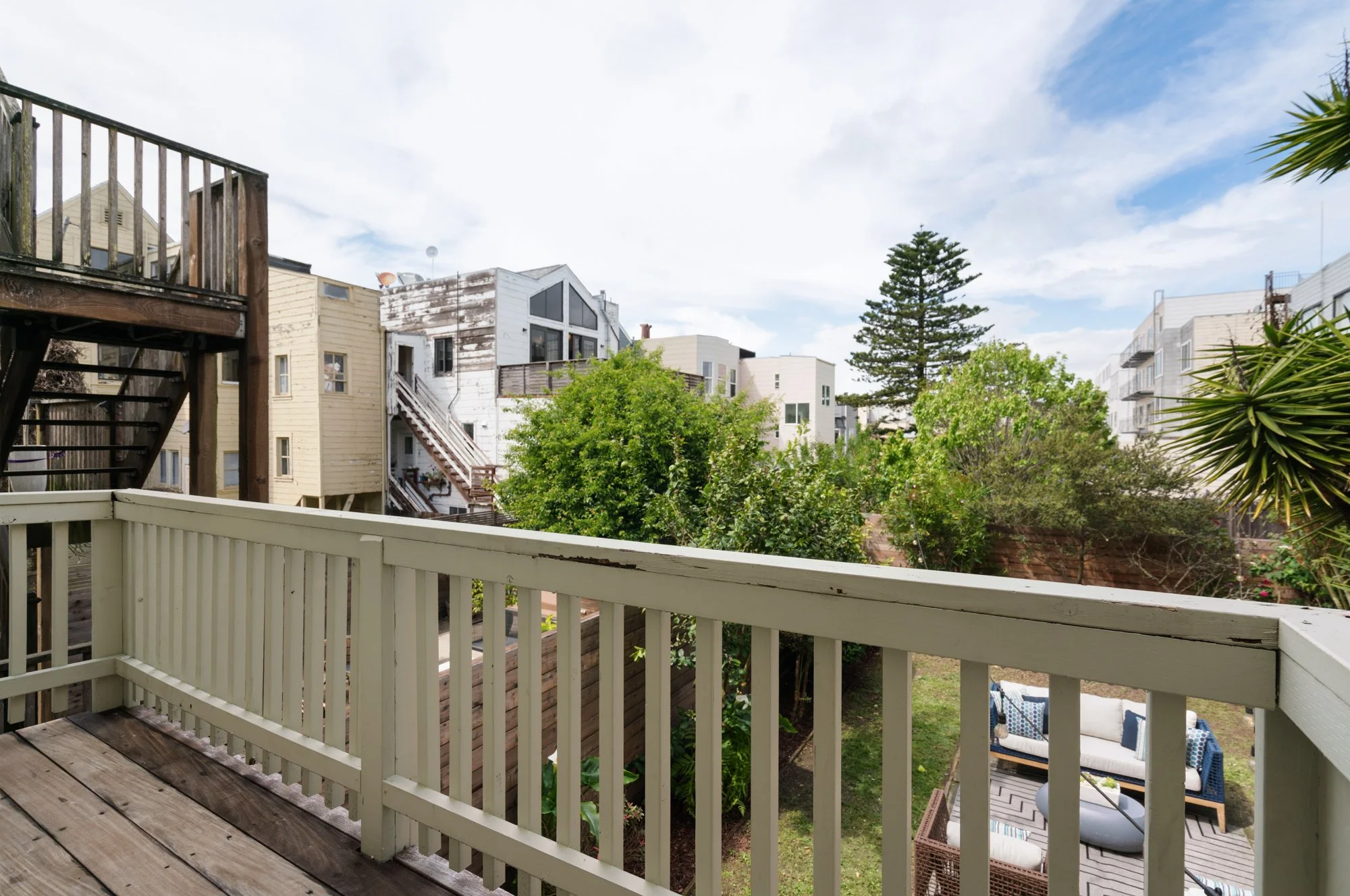 View from a balcony overlooking a backyard with outdoor seating, trees, and neighboring buildings under a partly cloudy sky.