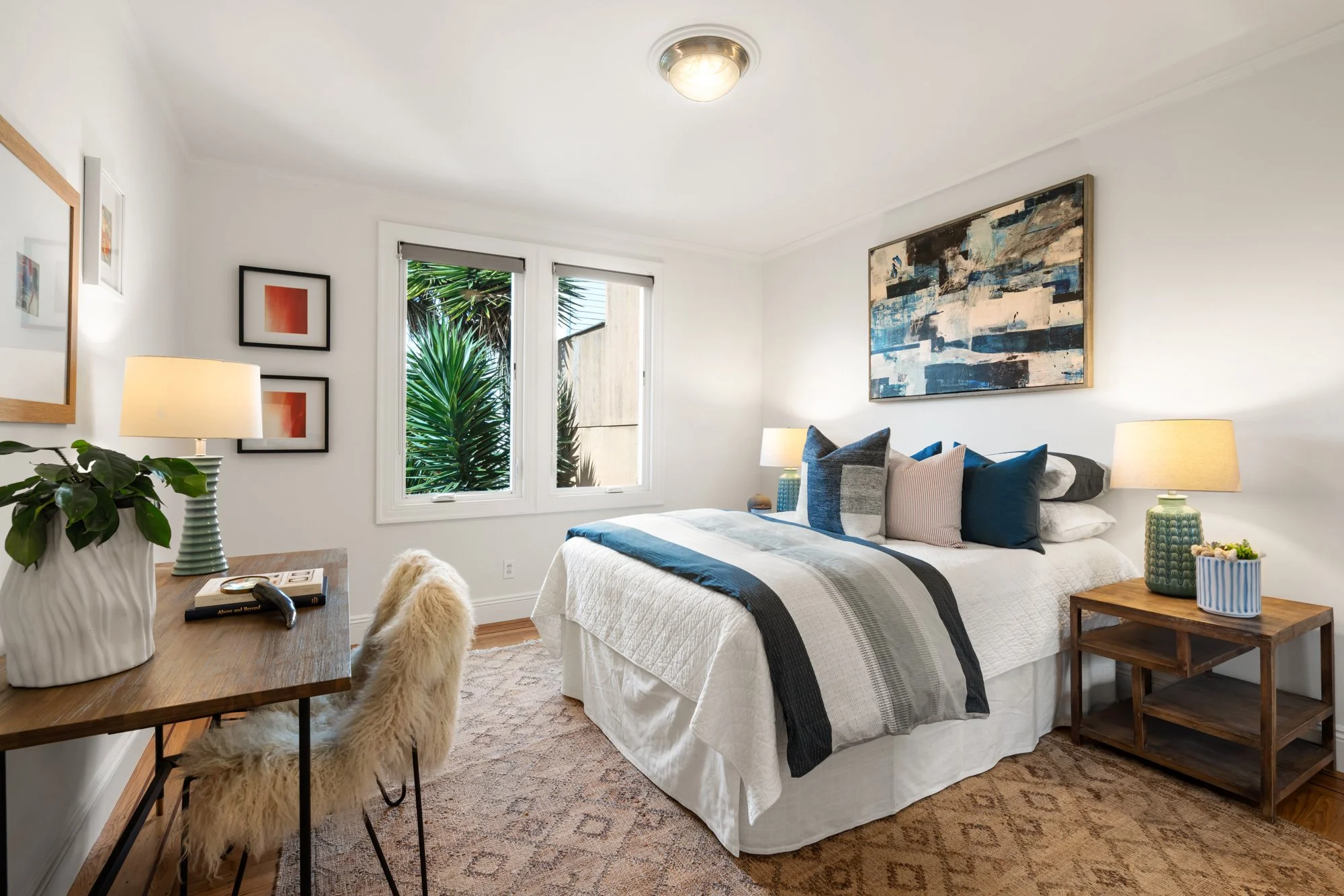 A cozy bedroom with a white bed, blue and gray pillows, two side lamps on wooden nightstands, a window with greenery outside, and artwork on the walls. There is also a wooden desk with a plant and a furry chair.