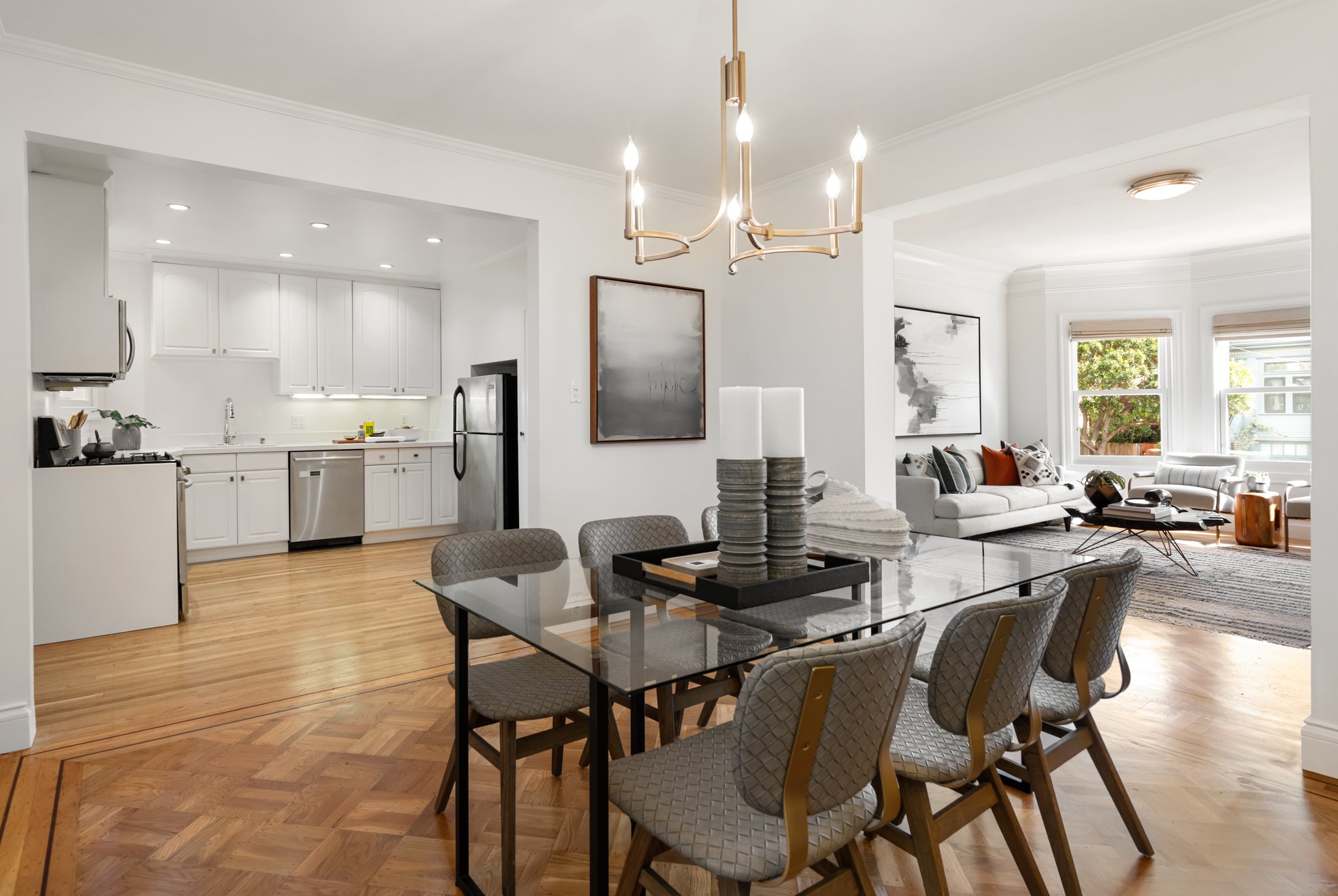 Modern open-concept living and dining area with white walls, hardwood floors, a dining table with six upholstered chairs, a chandelier, a sitting area with white sofas, a large window, and a kitchen with white cabinets and stainless steel appliances.
