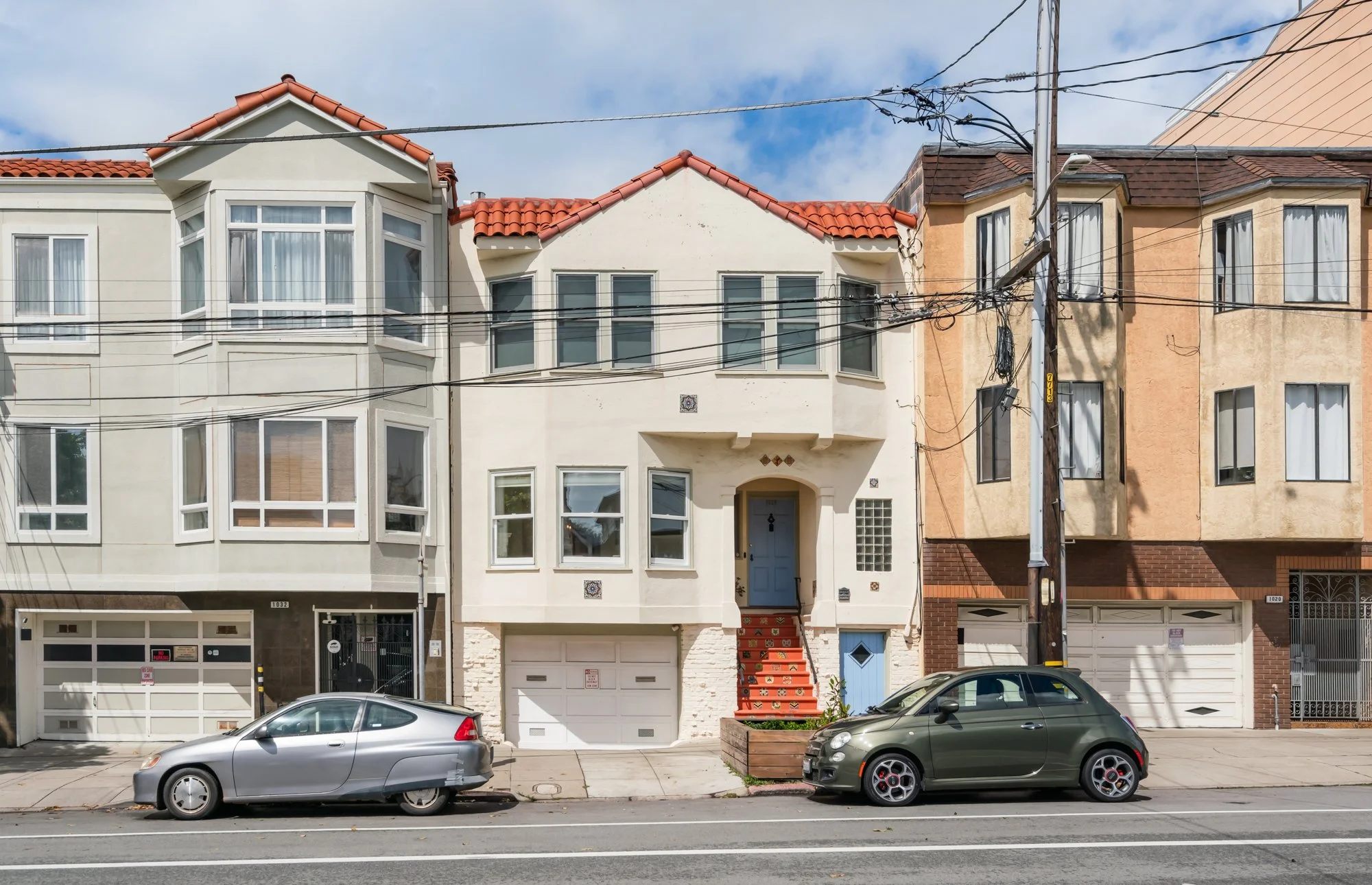 Row of three houses with cars parked in front, featuring distinct architectural styles and colors, under a partly cloudy sky.