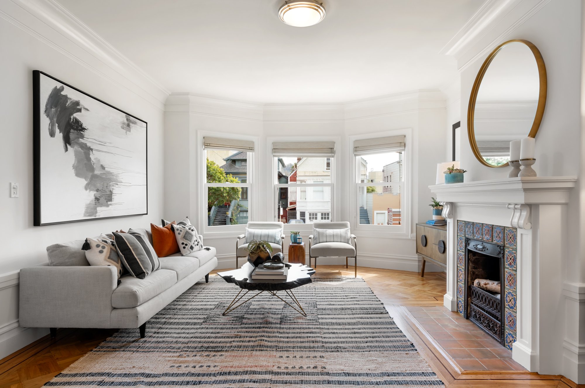 Bright living room with white walls, three windows, white sofa with throw pillows, black and white striped rug, black coffee table, two white armchairs, fireplace with colorful tiles, framed artwork, and gold-framed mirror on mantel