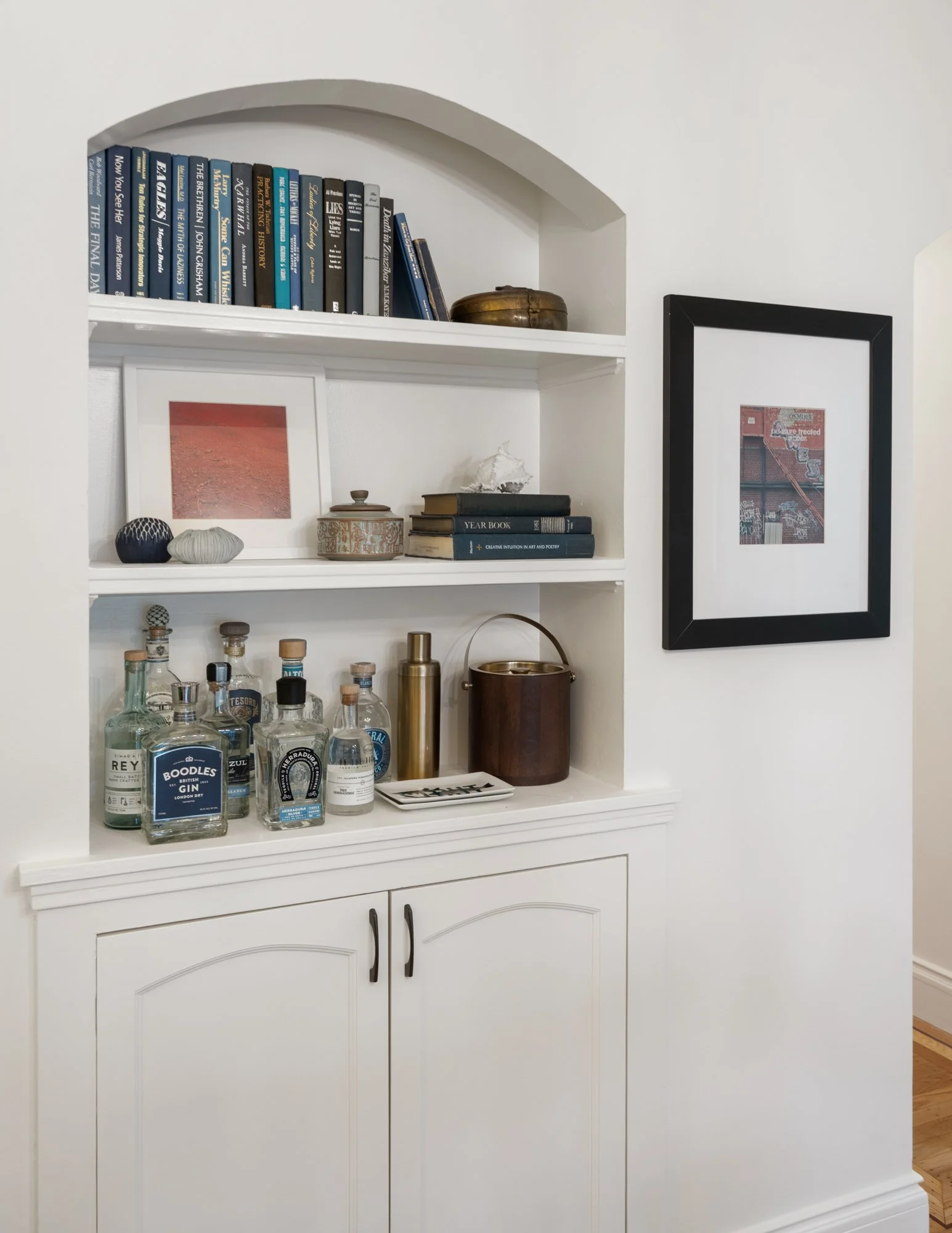 A white built-in shelf with three compartments holding books, decorative objects, and liquor bottles, with framed artwork on the wall nearby.