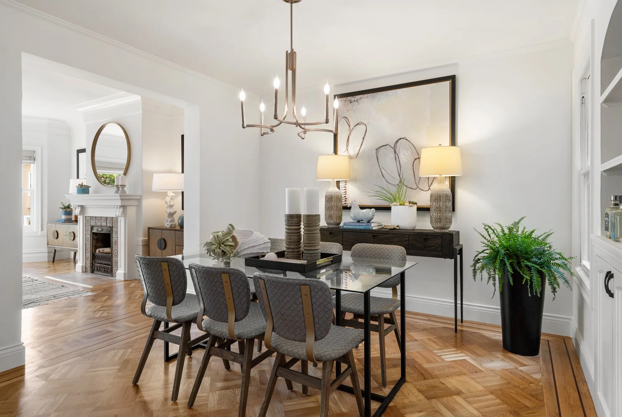 Dining room with a glass table, six patterned chairs, a dark sideboard with lamps and artwork, a chandelier, and a large potted fern.