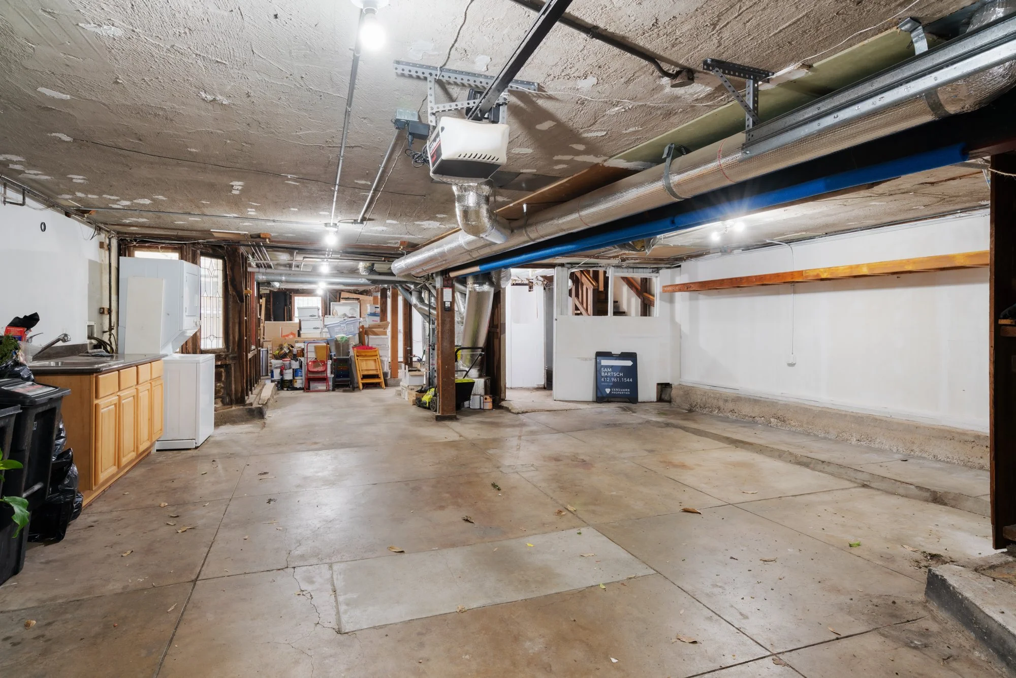 Empty garage with concrete floor, storage items at the back, clutter, exposed pipes on ceiling, and a small sink and cabinet on the left.