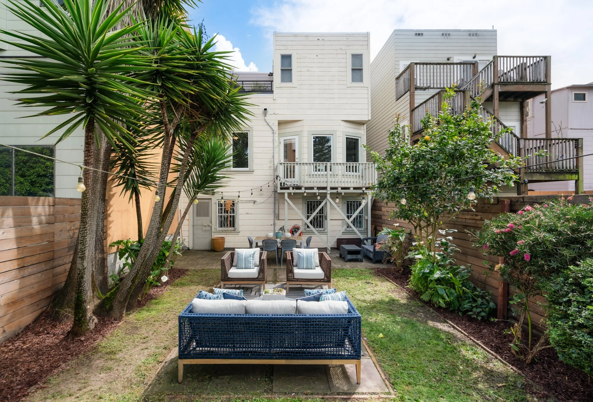 Backyard patio area with outdoor furniture, including a blue couch, six chairs with cushions, and some side tables, surrounded by a wooden fence, trees, and flowering bushes, with a white multi-story house in the background.
