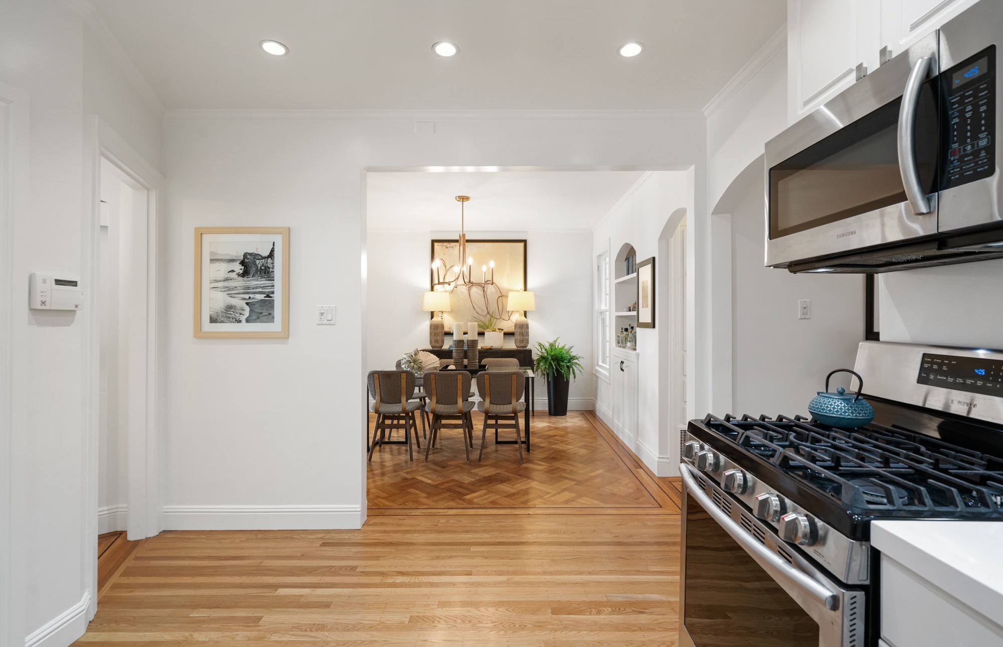 View of a bright, white kitchen with stainless steel appliances, including a microwave and a stovetop, leading into a dining area with a wooden floor and a chandelier.