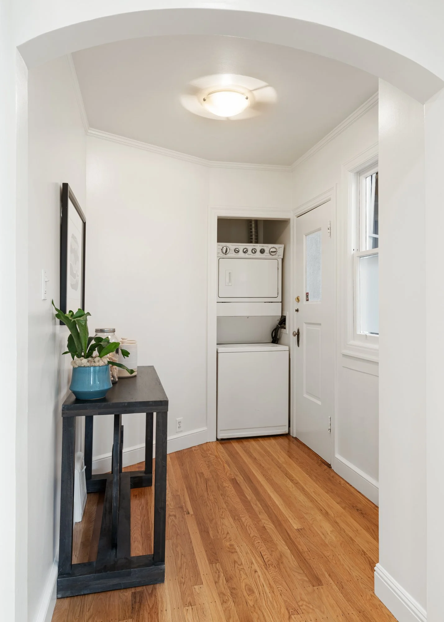 A small laundry area with a stacked washer and dryer, white walls, hardwood floor, a window, and a narrow black table with a potted plant.