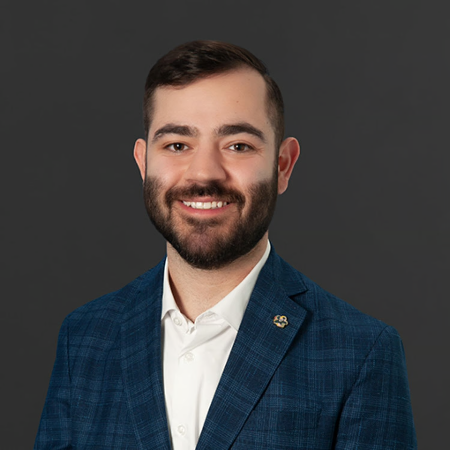 Portrait of a smiling young man with dark hair and beard, wearing a blue checked suit jacket and white shirt, against a dark gray background.