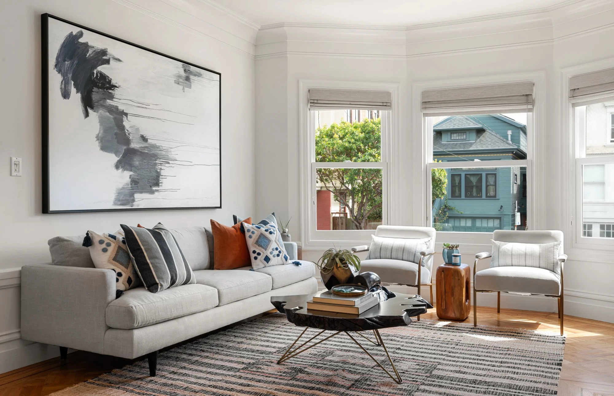 Bright living room with a white sofa, decorative pillows, two white armchairs, a black coffee table with books, and large windows showing trees and neighboring houses.