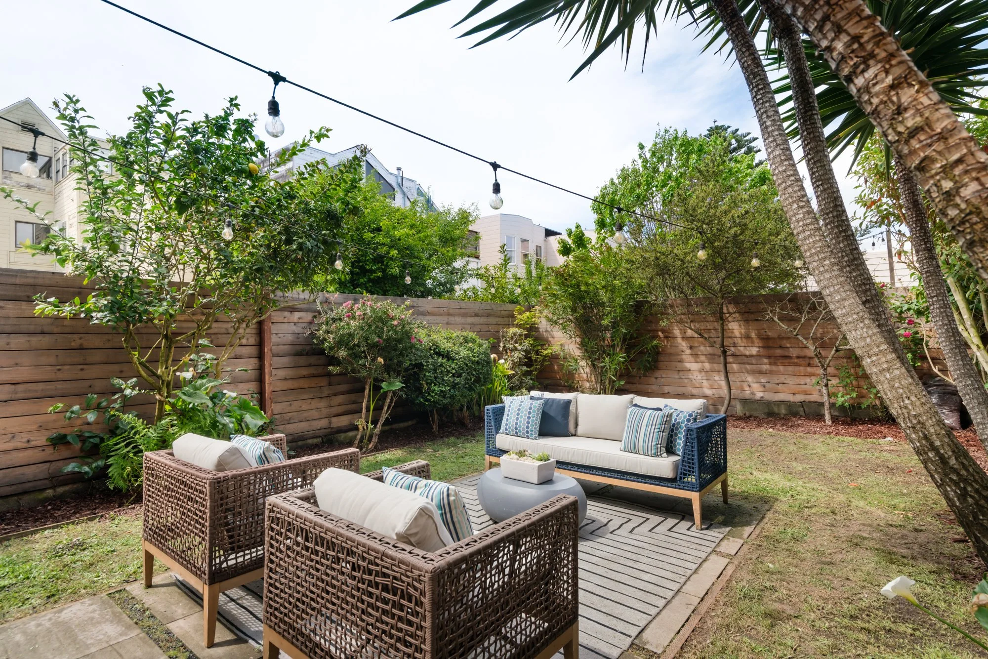 A cozy outdoor patio in a backyard with a wooden fence, string lights, green trees, and modern furniture including a cushioned sofa, armchairs, and a central coffee table, set on a striped outdoor rug.