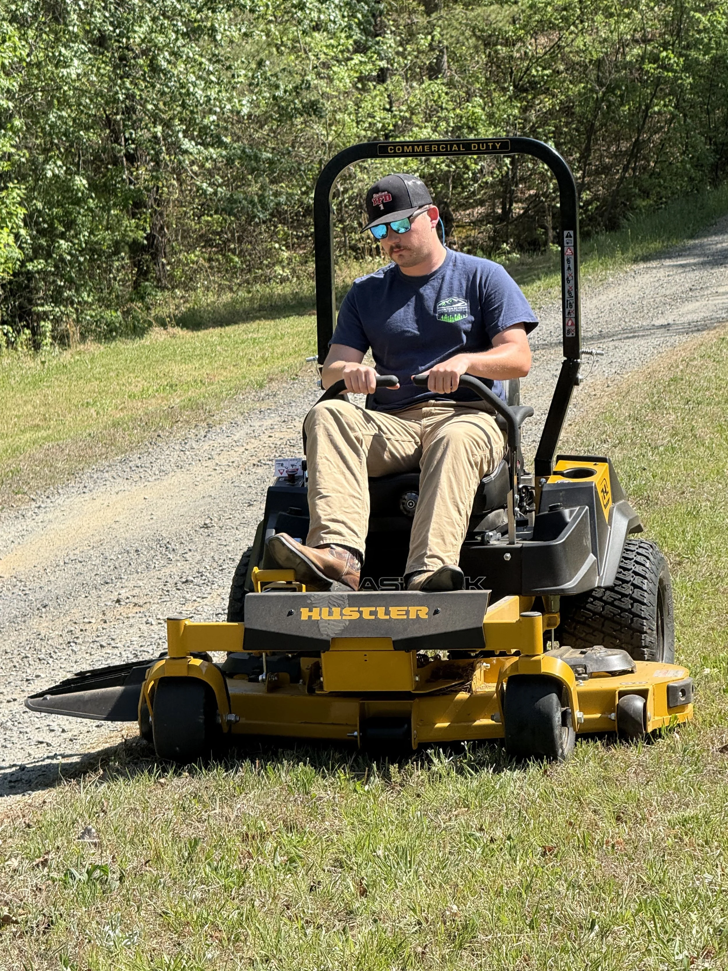 A man riding a yellow Hustler zero-turn mower on a grassy patch beside a gravel path, surrounded by trees.