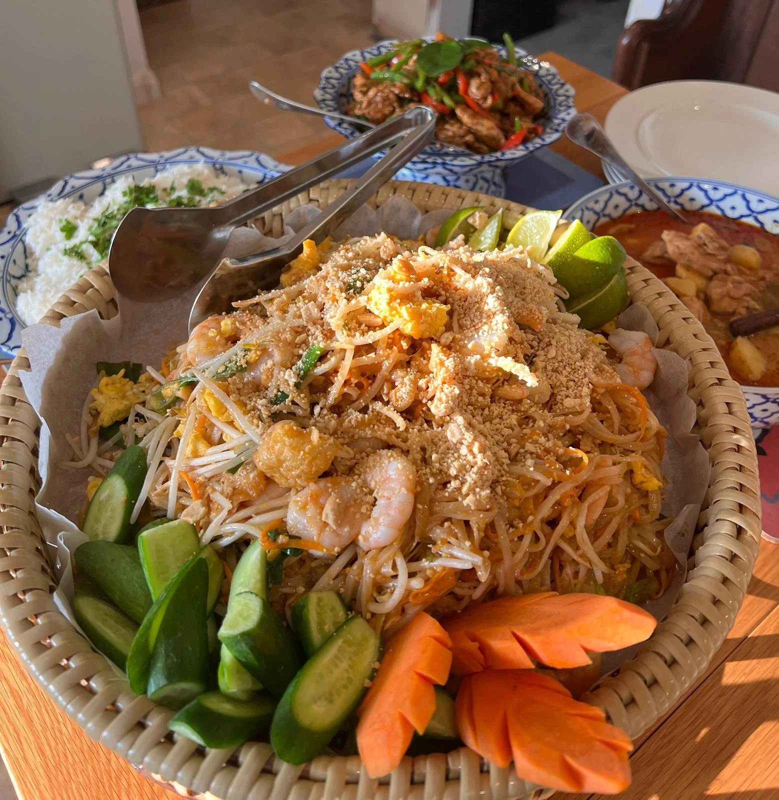 A basket of Thai-style seafood pad thai topped with crushed peanuts, served with lime wedges, cucumber slices, and orange carved flower decorations. In the background, there are bowls with stir-fried meat and vegetables and a bowl of chili shrimp.