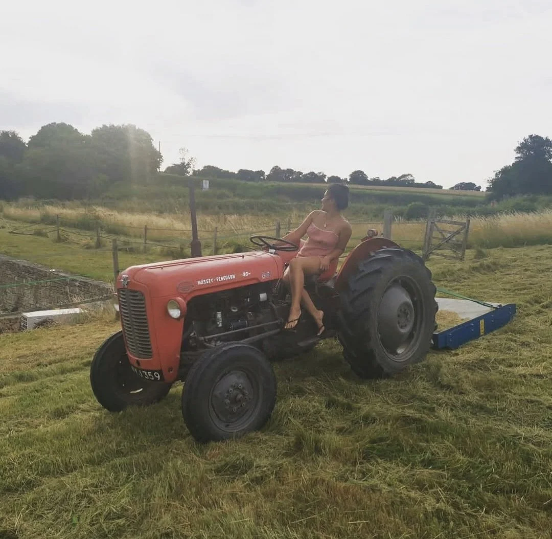 A woman sitting on a red Massey Ferguson tractor on a grassy field with open landscape and trees in the background.