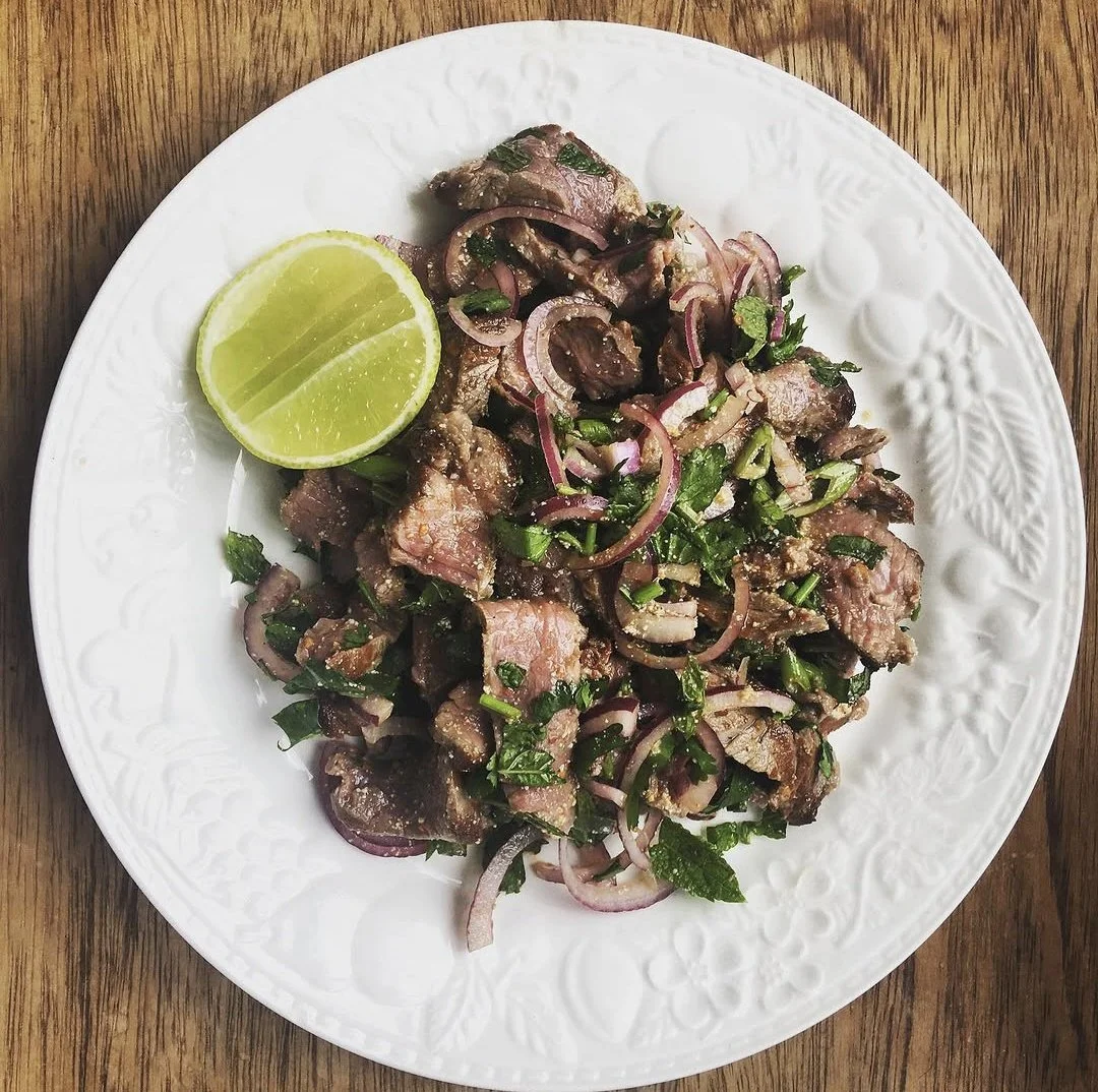 A white plate with an intricate pattern around the edge, holding chopped beef mixed with sliced red onions and chopped cilantro, garnished with a lime wedge, placed on a wooden surface.