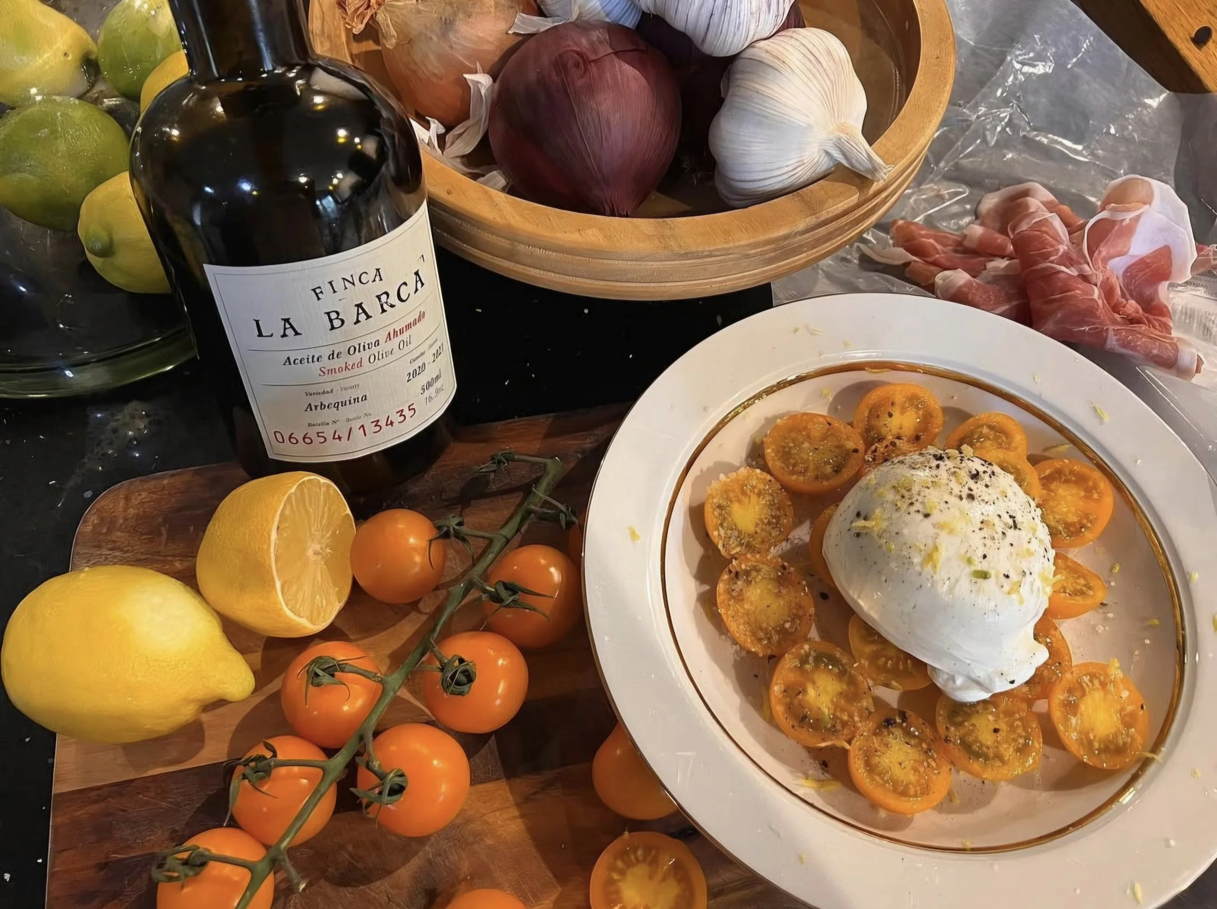 A variety of fresh produce and ingredients on a kitchen counter, including a bottle of smoked olive oil, yellow lemons, a bunch of orange tomatoes, red onions, garlic bulbs, sliced heirloom tomatoes, and a dish with poached eggs and roasted tomatoes.
