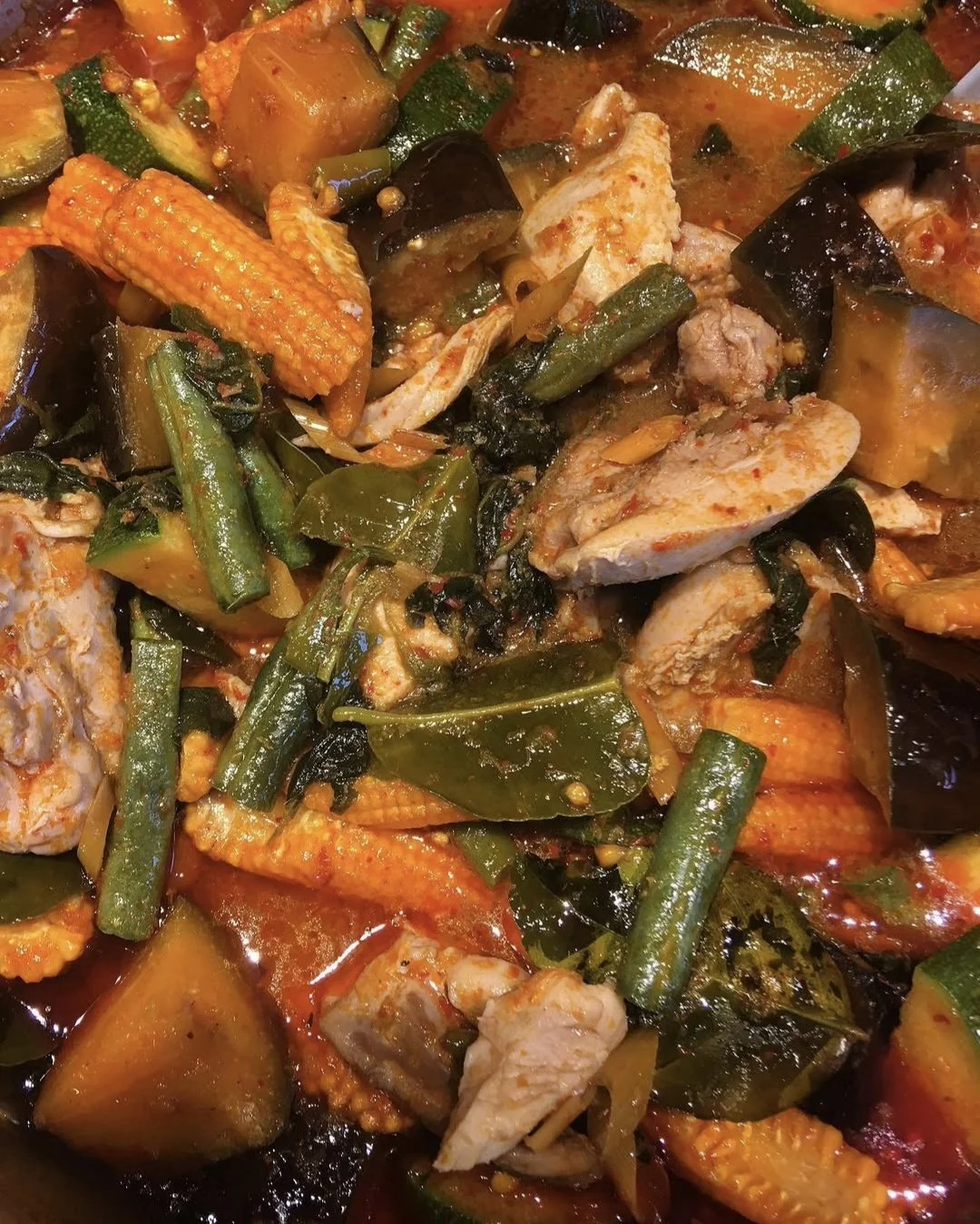 A close-up of a vegetable and chicken stew with green beans, corn, eggplant, zucchini, and chunks of chicken in a spicy, tomato-based sauce.