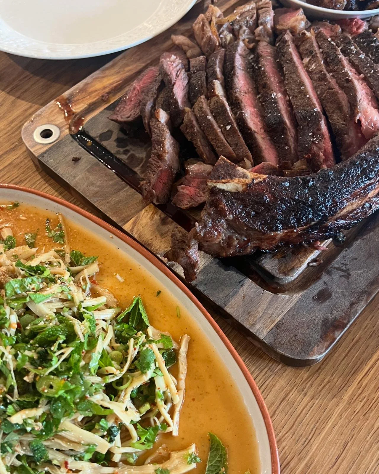 Sliced cooked steak on a wooden cutting board with a dark crust, surrounded by bowls of salad and other side dishes.