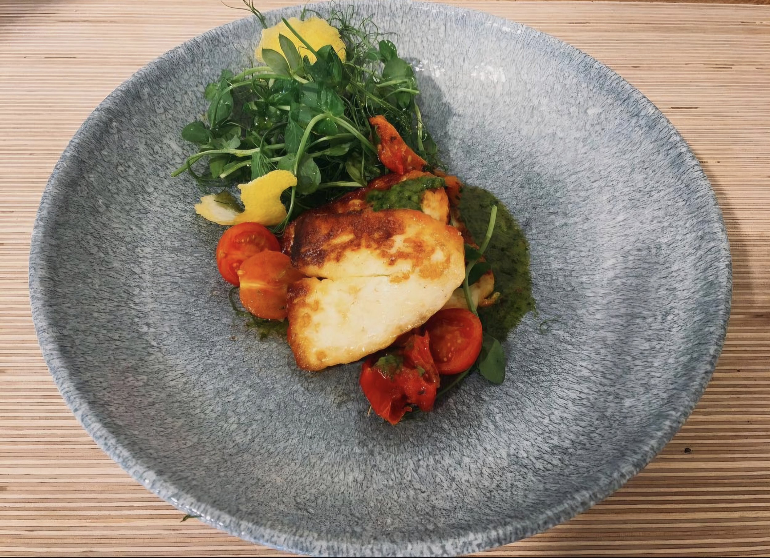 A plated dish with seared fish, cherry tomatoes, green sauce, and a side of fresh greens with lemon slices on a gray textured plate, on a wooden surface.