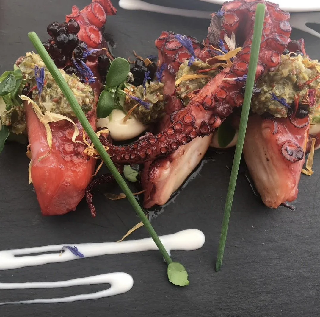 Grilled octopus tentacles topped with eggplant and garnished with edible flowers, microgreens, and drizzled with white sauce, served on a black slate plate.