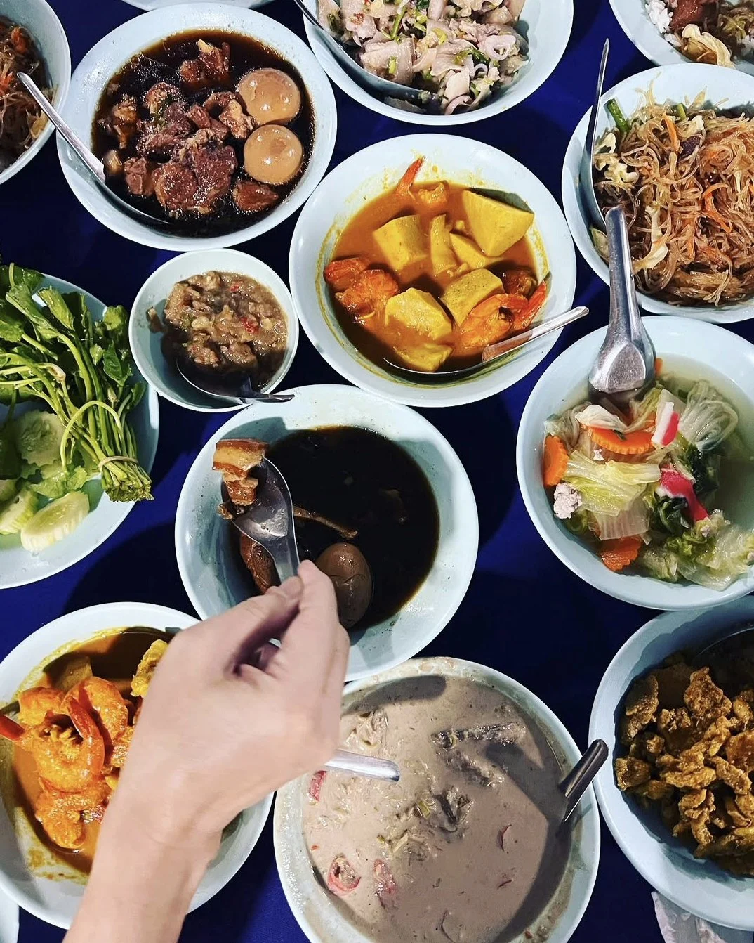 A variety of Asian dishes on a table, including stewed eggs, curries, salads, and soups, with a person serving themselves with a spoon.