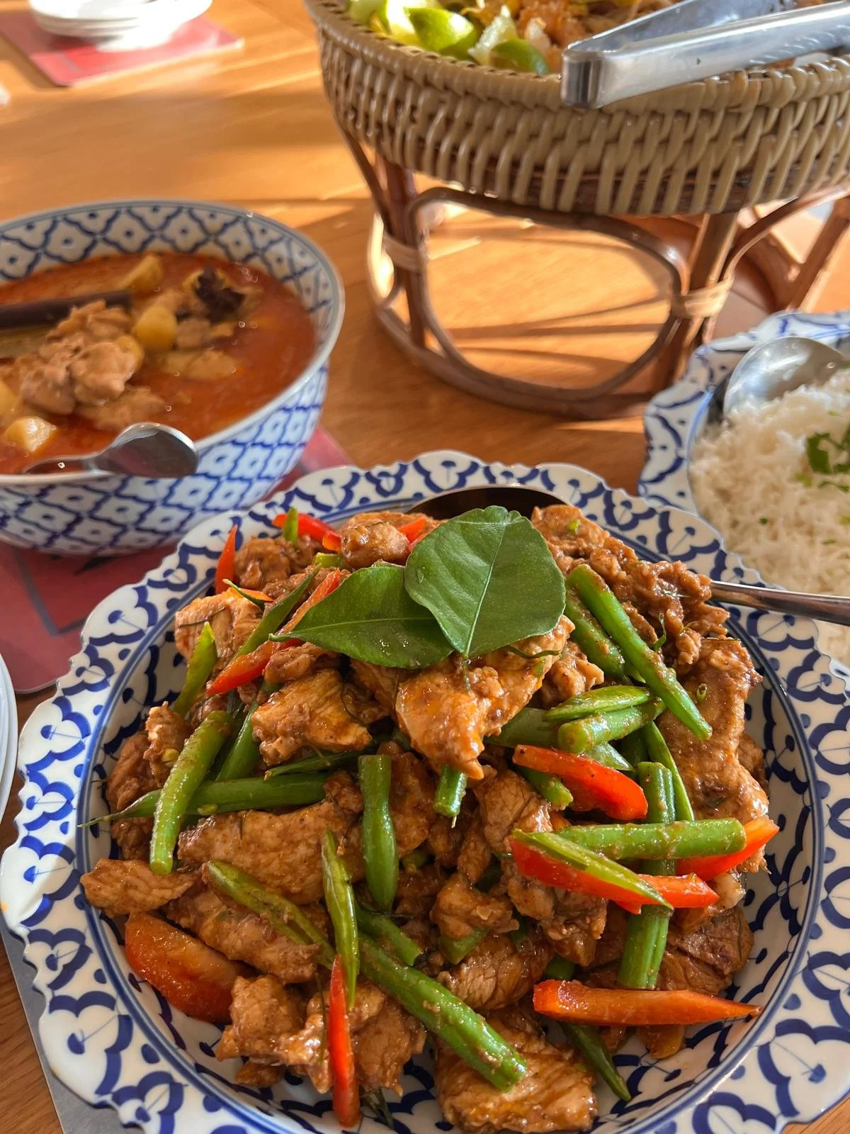 Close-up of a plate of stir-fried chicken with green beans and red bell peppers garnished with a bay leaf, accompanied by other dishes and a basket of rice in the background.
