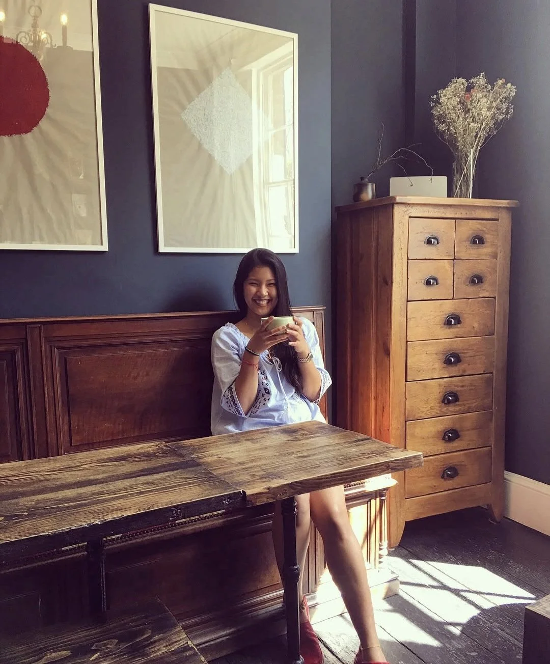 A woman with long dark hair sitting at a wooden table, smiling and holding a cup in a cozy room with dark blue walls, wooden furniture, and wall art.