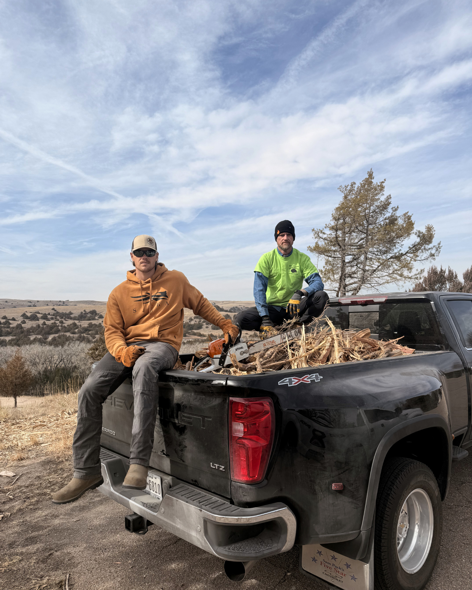 Two men are standing in a pickup truck filled with chopped wood in an open landscape. One man in a brown hoodie and sunglasses is sitting on the truck's edge while the other, wearing a bright green shirt, black gloves, and a black beanie, is kneeling inside the truck bed. A tree and a partly cloudy sky are visible in the background.