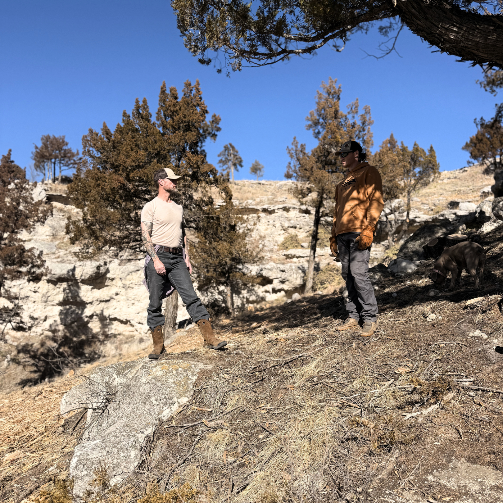 Two men stand on rocky terrain surrounded by trees under a blue sky. One man has a beard, tattoos, and wears a cap, t-shirt, jeans, and boots. The other man wears sunglasses, a baseball cap, a brown jacket, gloves, and gray pants. A dog is partially visible on the right side of the image.