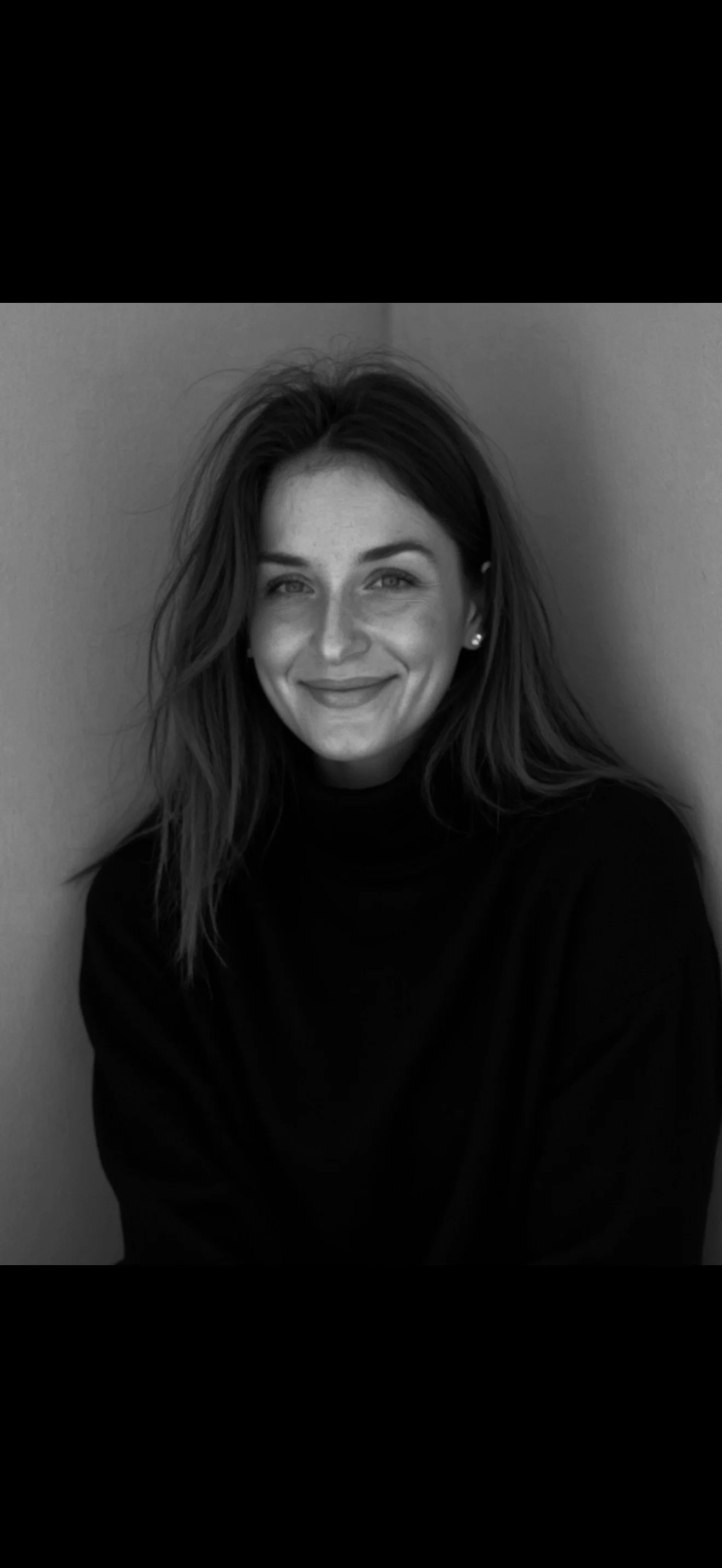 Une femme souriante à la peau claire, avec des cheveux mi-longs ondulés, portant un haut noir, posant contre un mur. L'image est en noir et blanc.