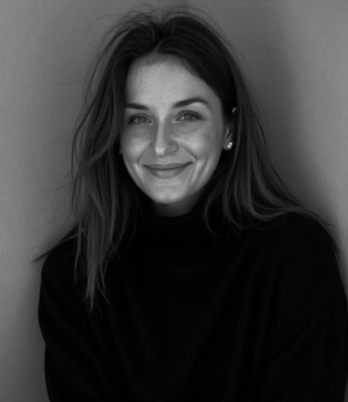 Portrait en noir et blanc d'une femme souriante avec des cheveux longs et ondulés, portant un pull noir, assise contre un mur